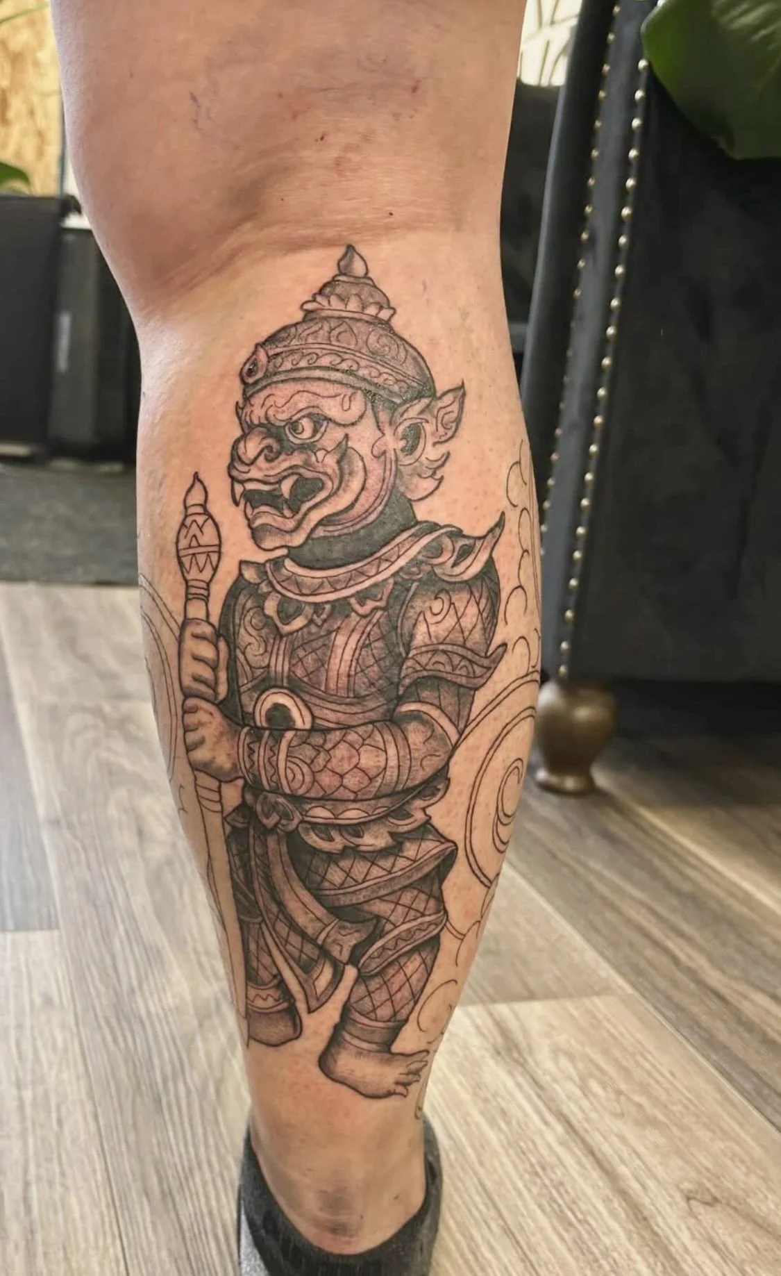 Tattoo of a mythological creature resembling a guardian figure, holding a staff, with traditional armor and detailed ornamentation, on a person's calf.