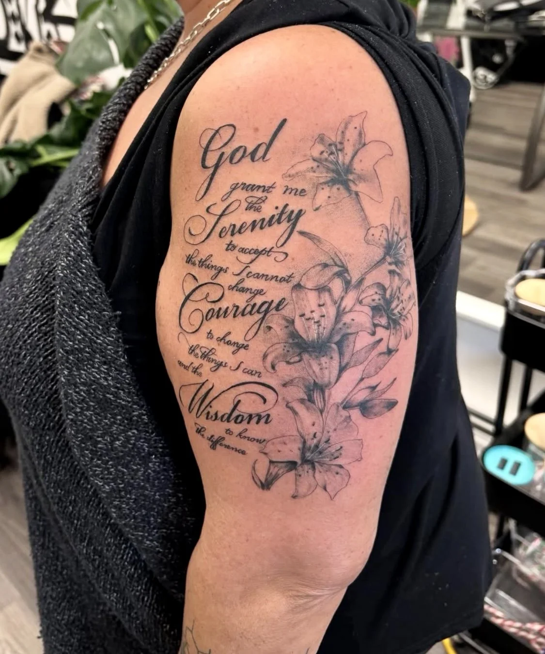 A tattoo on a person's upper arm features a quote about serenity, courage, and wisdom, intertwined with a detailed floral design of lilies.