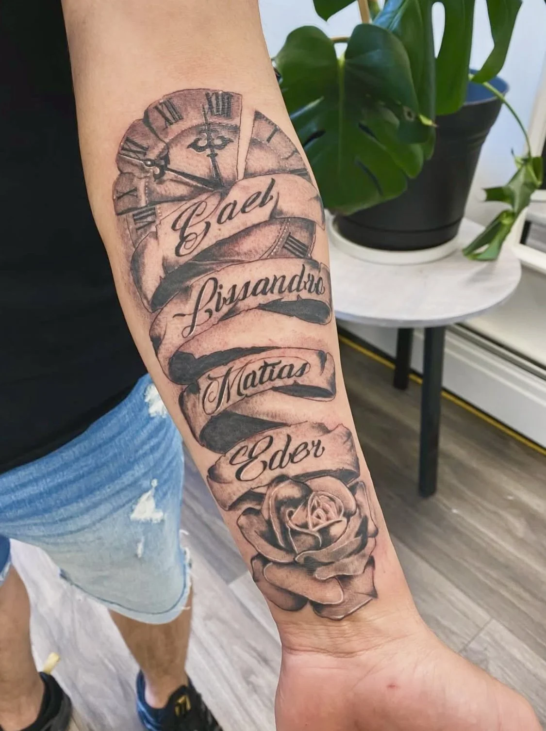 Tattoo on person's arm featuring a clock and a rose, with words 'Gael', 'Lissandro', 'Matias', and 'Eder' written on banners.