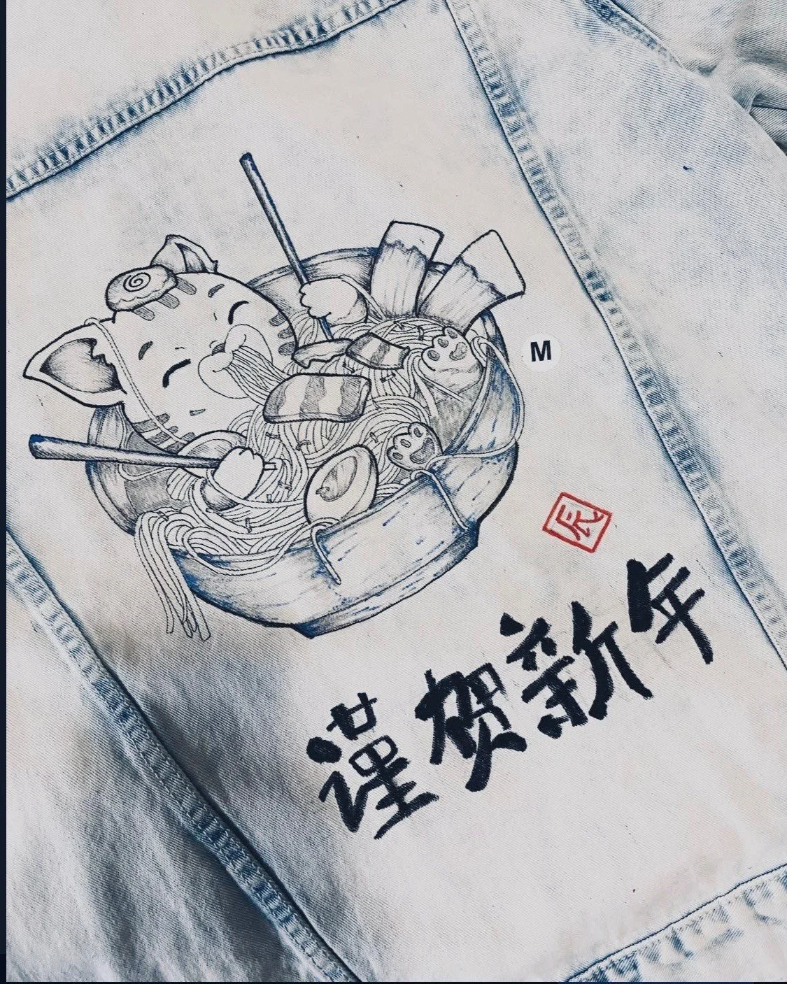 Embroidery of a bowl of ramen with a cute cat face, chopsticks, and various toppings, on light-colored fabric with Chinese characters and a red stamp.