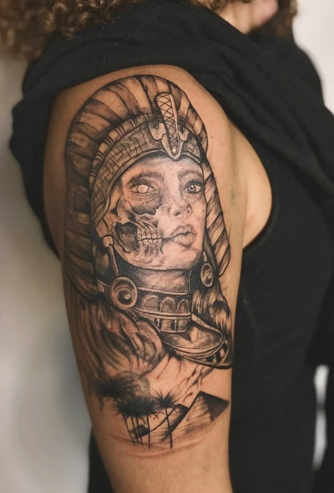 Tattoo of a woman with a headdress, blending her face with a skull, on an upper arm.