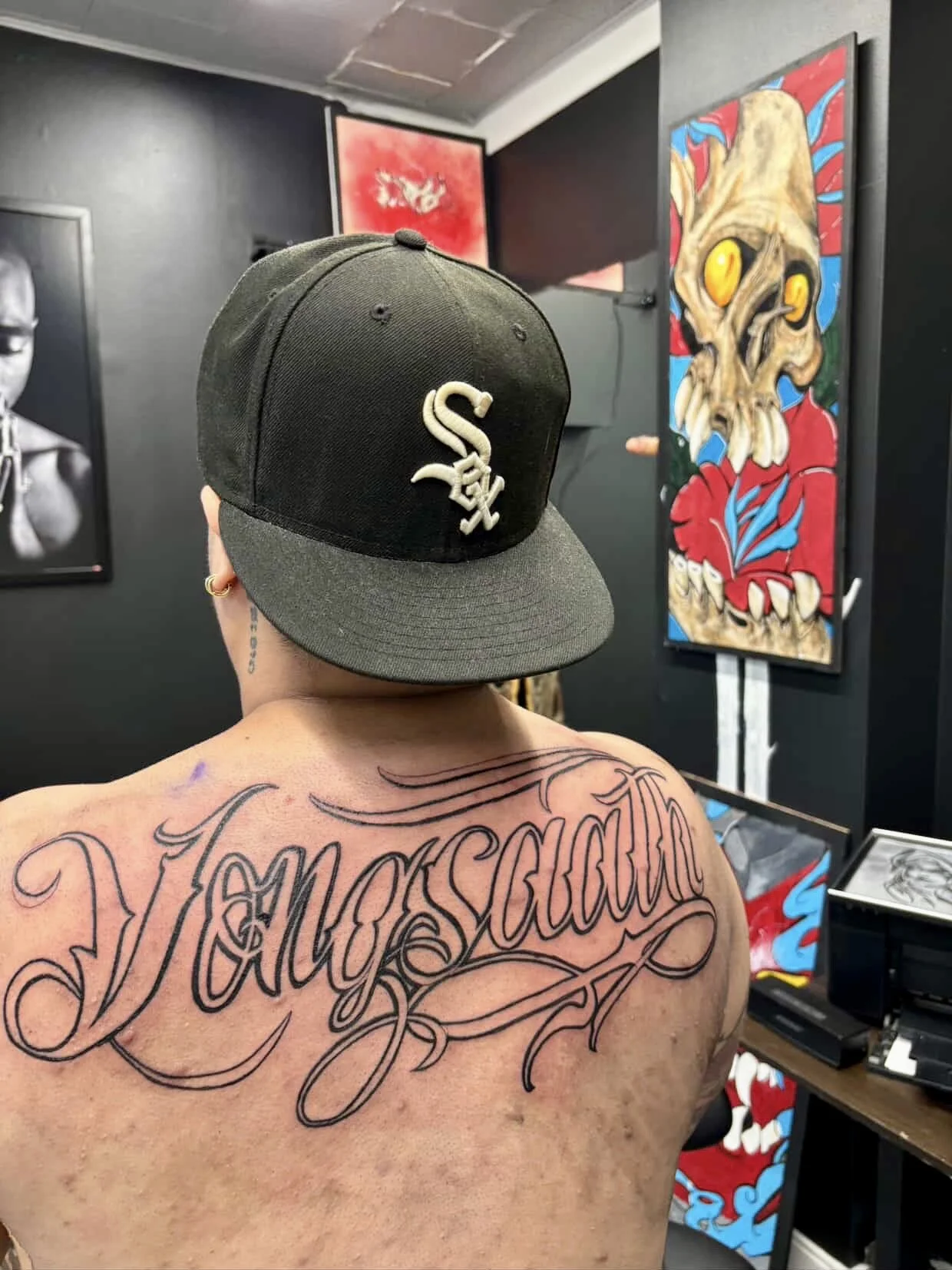 A person with a tattoo that says 'Tayronsain' on their back, wearing a black baseball cap with a white emblem, in an art studio with paintings on the wall.