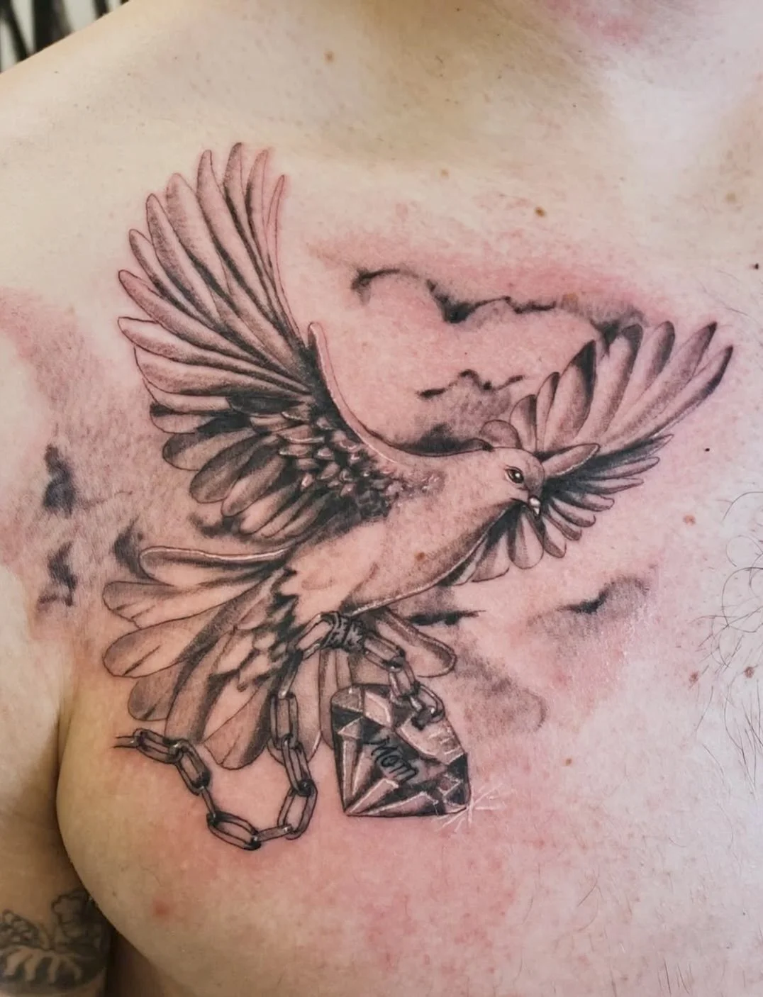 Black and gray tattoo of a bird flying with a chain and a gemstone pendant hanging from its beak, on a person's chest.