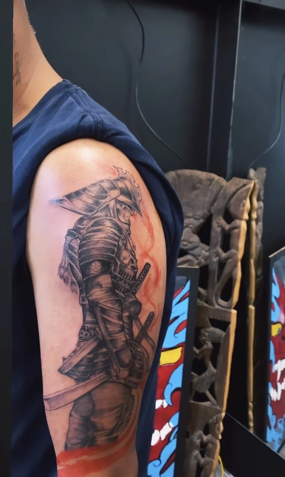 Tattoo of a samurai warrior in armor with a helmet and two swords on the arm of a person wearing a dark sleeveless shirt.