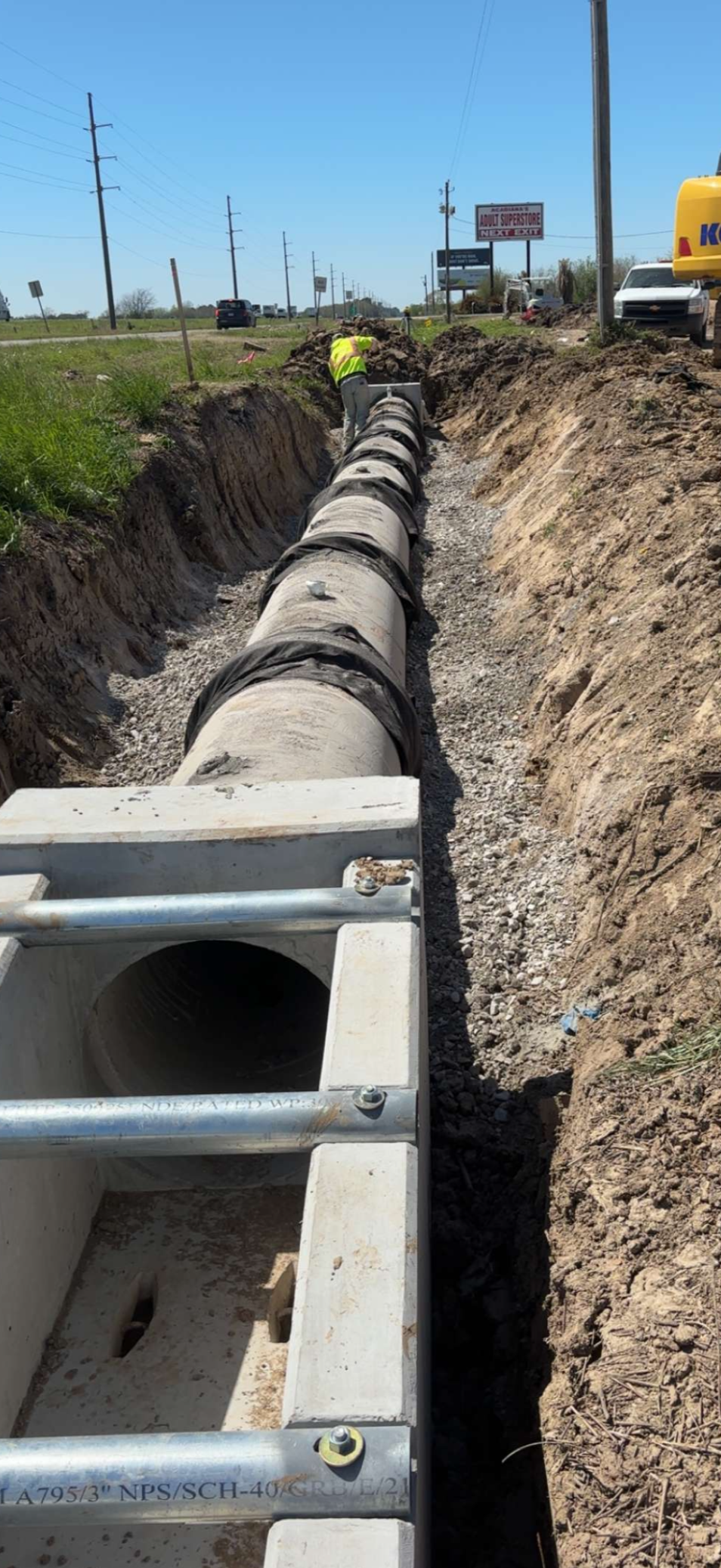 Construction worker installing large concrete storm drain pipe in a dug trench along a roadside with power lines and vehicles in the background.