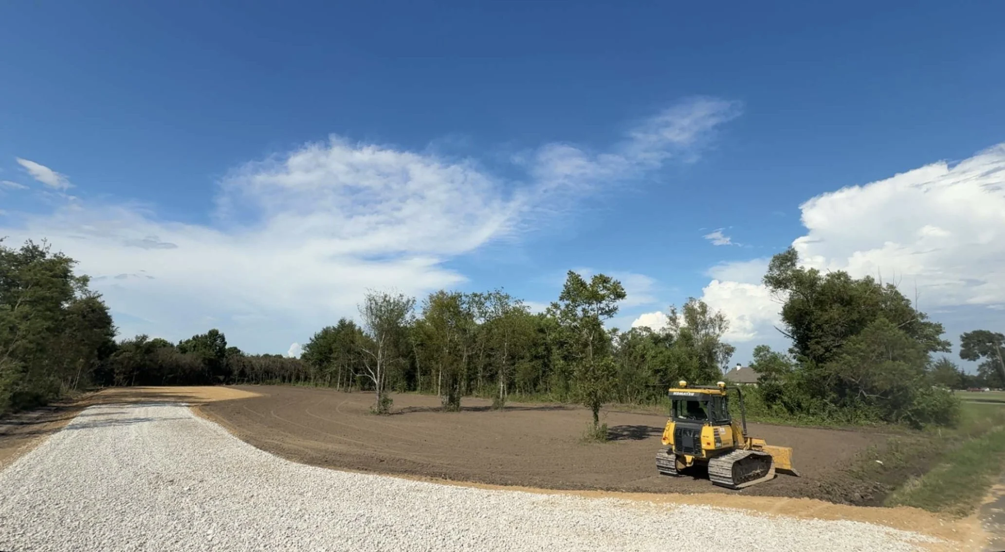 A construction site with a yellow bulldozer on a dirt surface, a gravel pathway, and trees in the background under a blue sky with scattered clouds.
