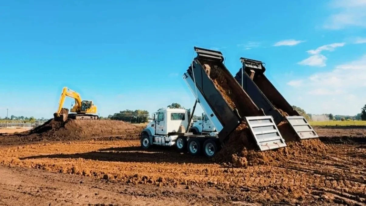 Construction site with a yellow excavator on a dirt mound and a white dump truck unloading soil.