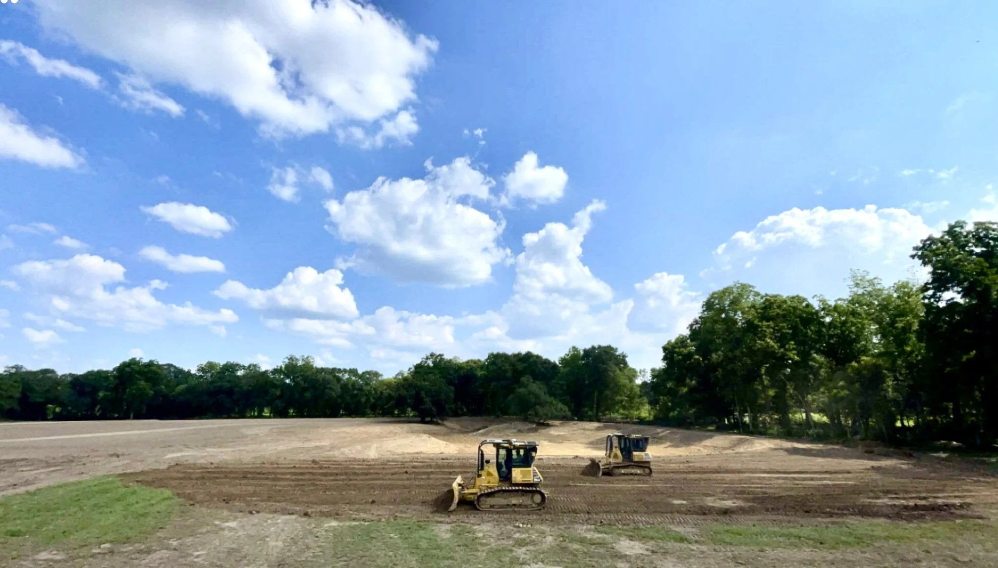 Construction site with bulldozers working on leveling the ground surrounded by green trees under a blue sky with scattered clouds.