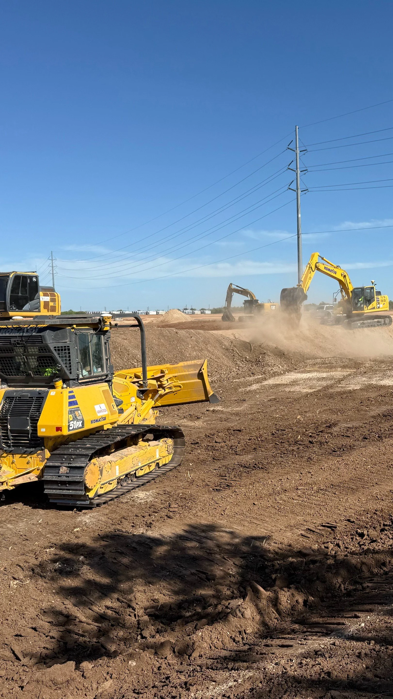 Construction site with yellow bulldozers and excavators moving dirt under power lines and clear blue sky.