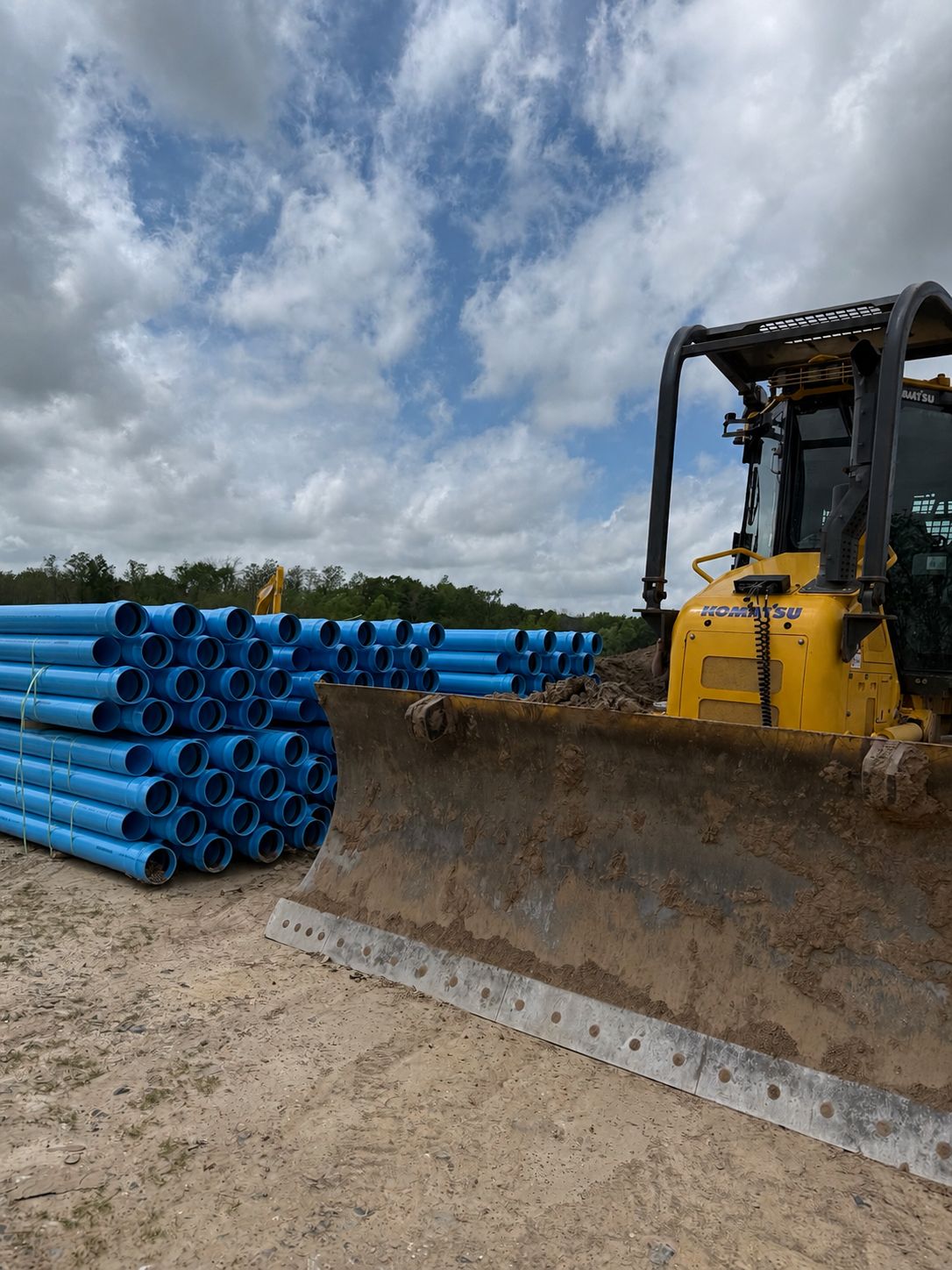 Construction site with a yellow bulldozer and a large stack of blue pipes under a partly cloudy sky.