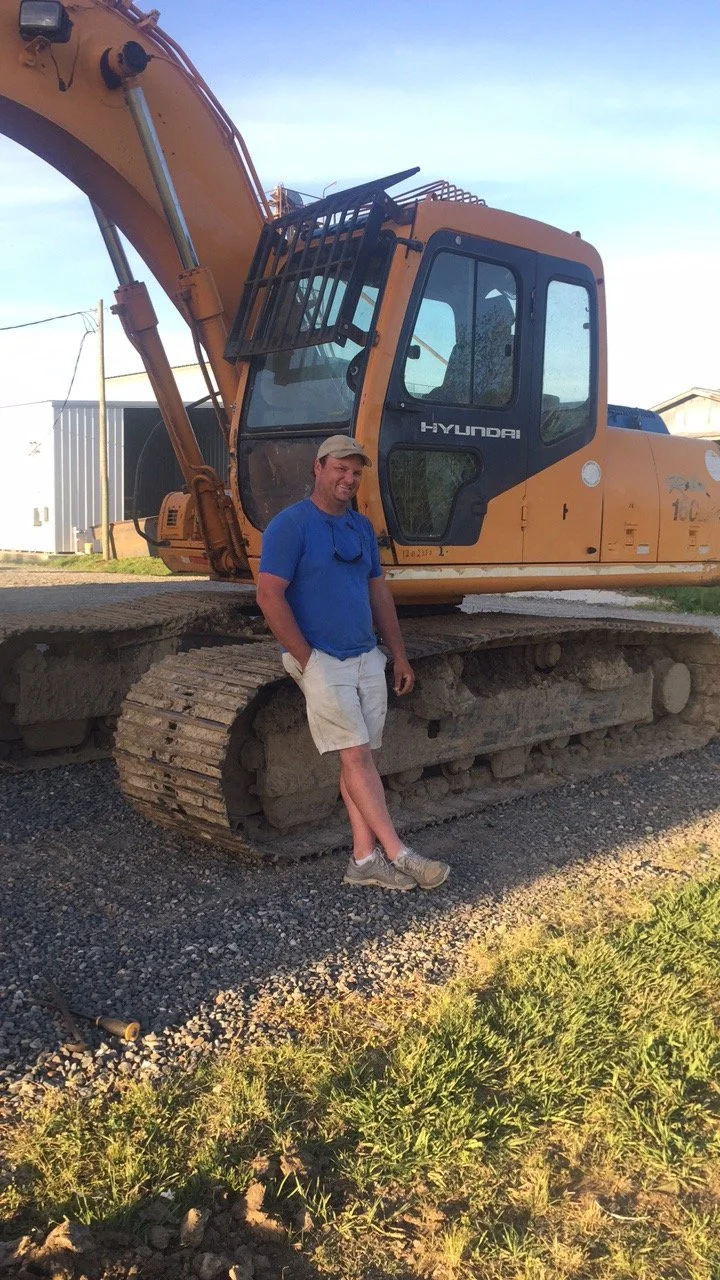 A man wearing a blue shirt, khaki shorts, and a beige cap standing in front of a yellow Hyundai excavator with black tracks outdoors during daytime.