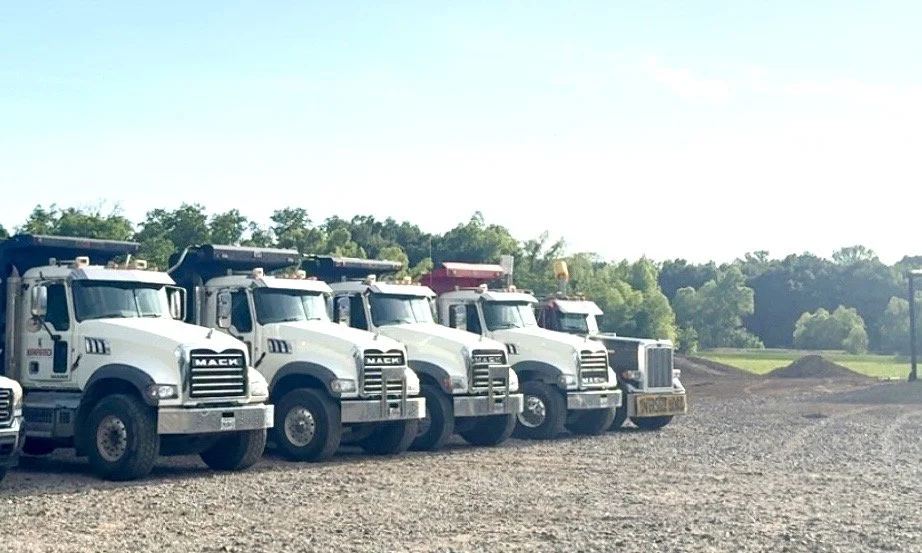 A row of six white dump trucks parked on a dirt lot with green trees and a blue sky in the background.