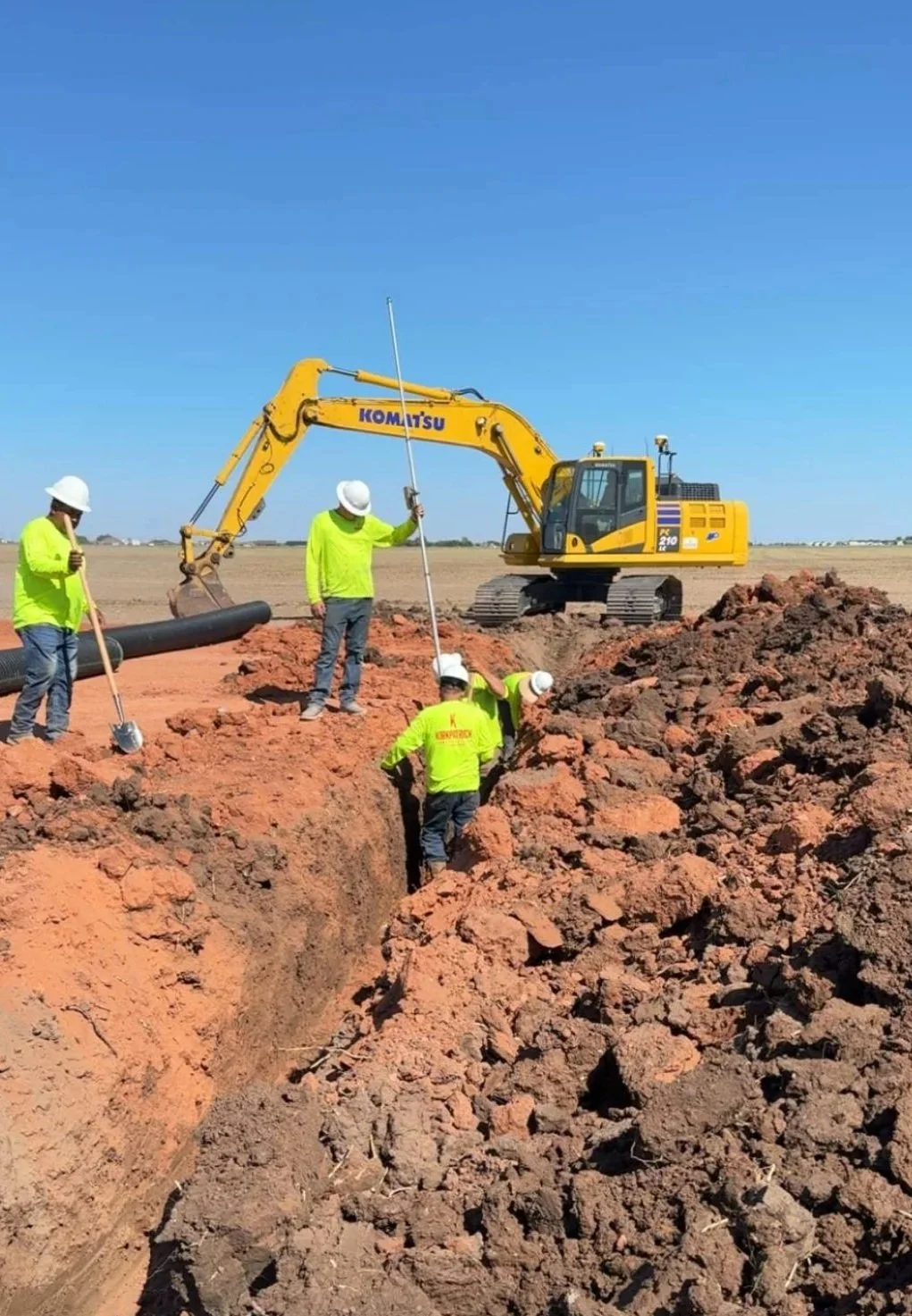 Construction workers in yellow safety vests and white hard hats work on a trench at a construction site with a yellow excavator in the background, under a clear blue sky.
