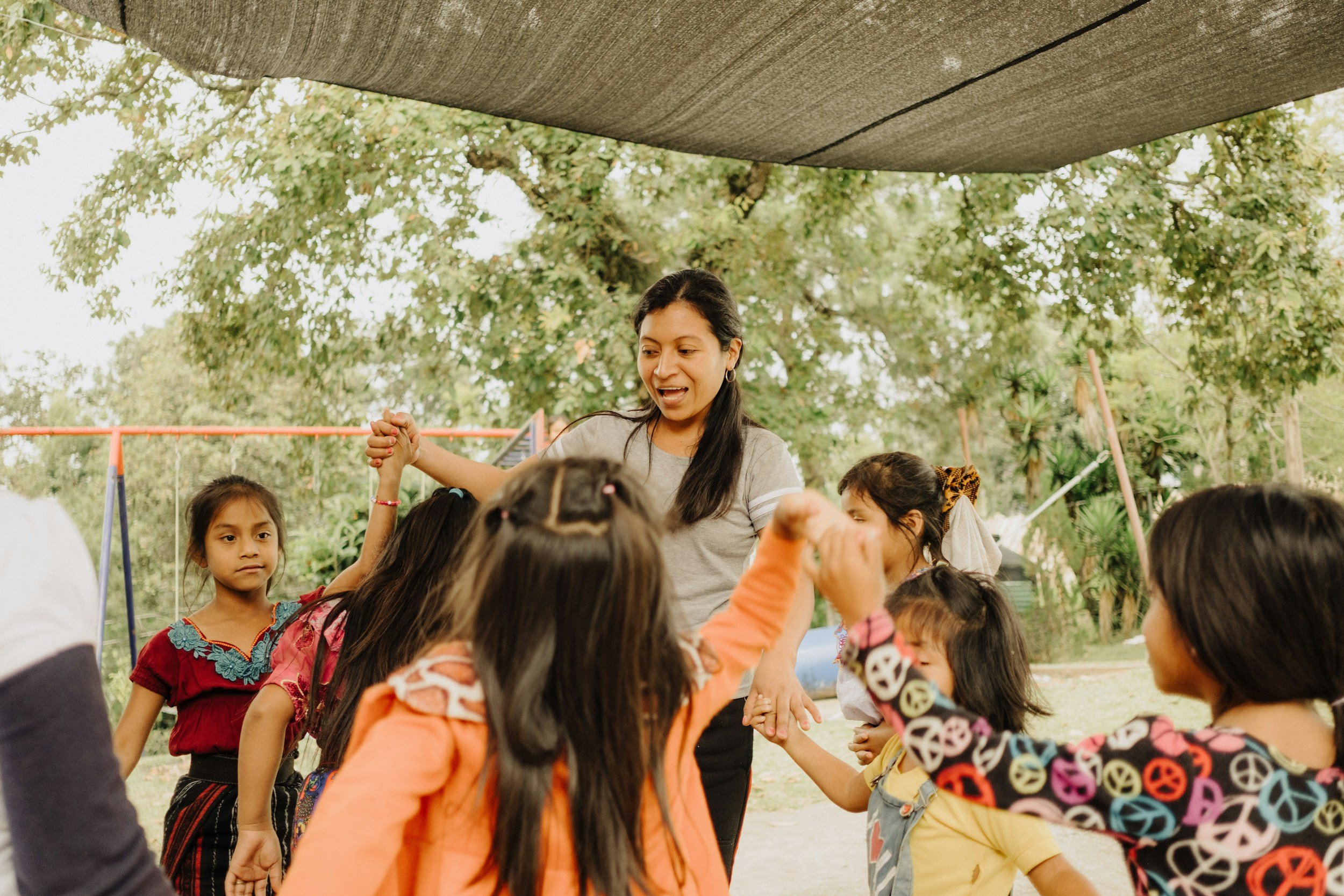 A group of children and an adult woman holding hands and playing together outdoors under a tree canopy.