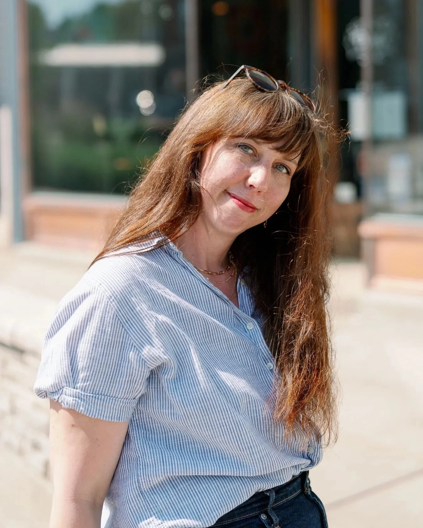 A woman with long red hair, blue eyes, and fair skin smiling outdoors. She is wearing a light blue and white striped shirt, black sunglasses on her head, and a gold necklace. The background shows a blurred building and greenery.