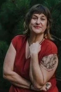 Woman with short brown hair and tattoos on her arm, wearing a red top, outdoors with trees in the background, smiling with her hand on her shoulder.