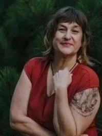 A smiling woman with shoulder-length brown hair and bangs, wearing a red top, posing outdoors with greenery in the background.