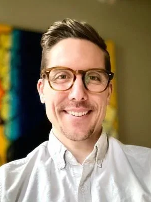 A man with dark hair and glasses smiling at the camera, wearing a white collared shirt.
