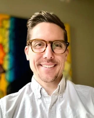 Jacob Moon, wearing a white button-up shirt, posing indoors with a colorful abstract artwork in the background.