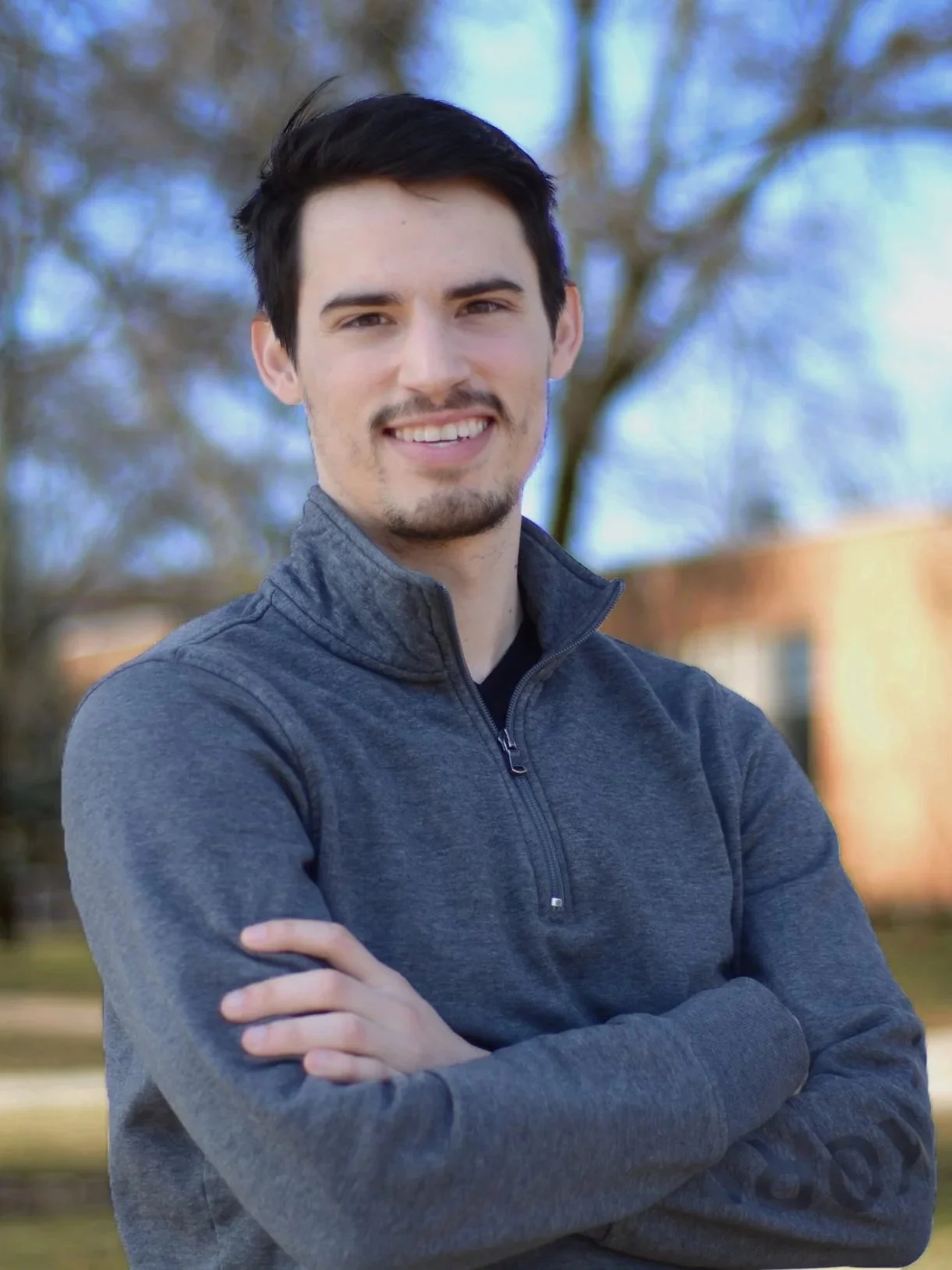 A young man with dark hair and a goatee smiling outdoors, wearing a gray zip-up jacket with arms crossed, with trees and a building in the background.