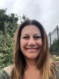 Smiling woman with long light brown hair outdoors, with trees and a fenced area in the background.
