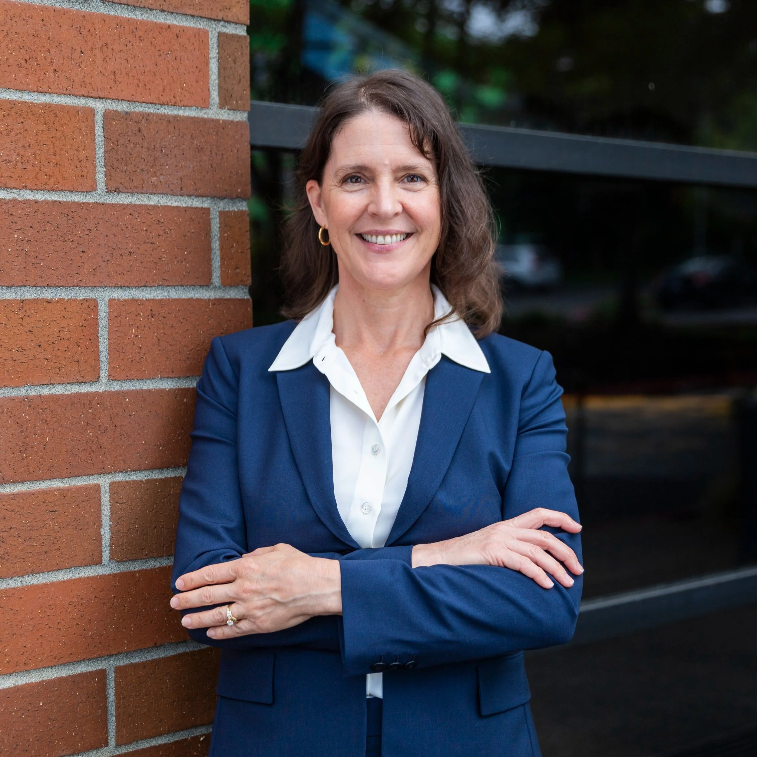 A woman in a navy blazer and white shirt standing outdoors against a brick wall, smiling with arms crossed.