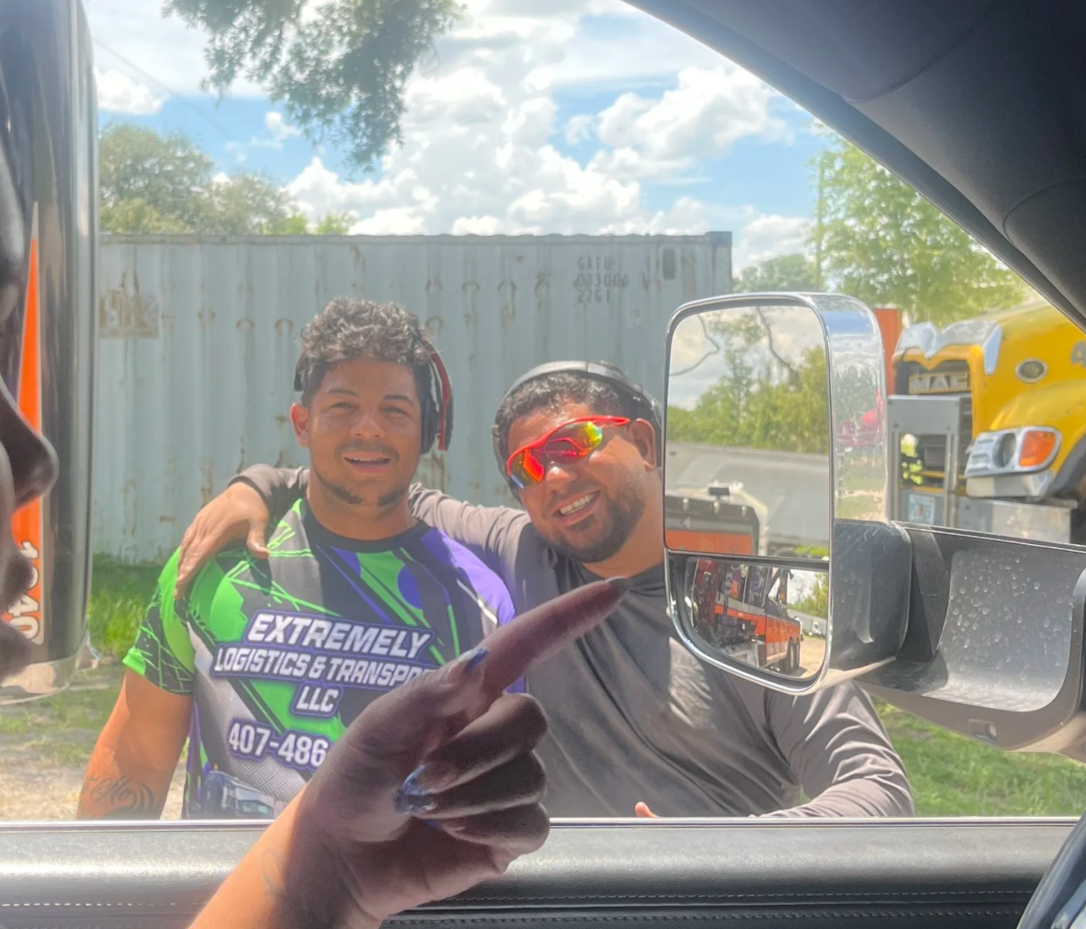 Two men standing outside, smiling and posing for a selfie taken from inside a vehicle. One man is wearing sunglasses and the other is wearing a shirt that says 'Extremely Logistics & Transport LLC.' Background features a shipping container, a yellow truck, and a partly cloudy sky.