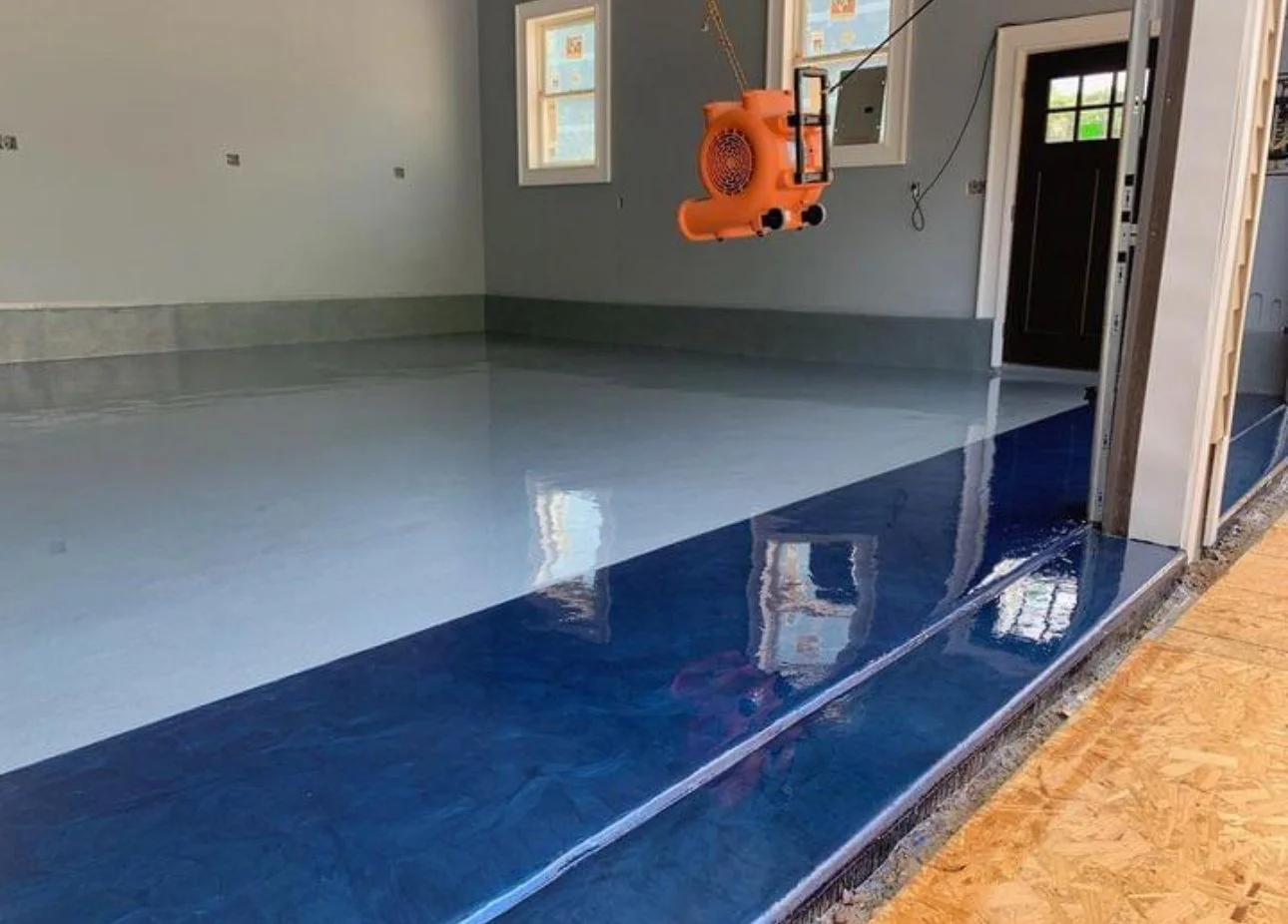 Newly finished interior space with a glossy light blue and dark blue epoxy floor, gray walls, multiple small windows, and an orange dust extraction machine hanging from the ceiling, indicating ongoing construction or renovation work.