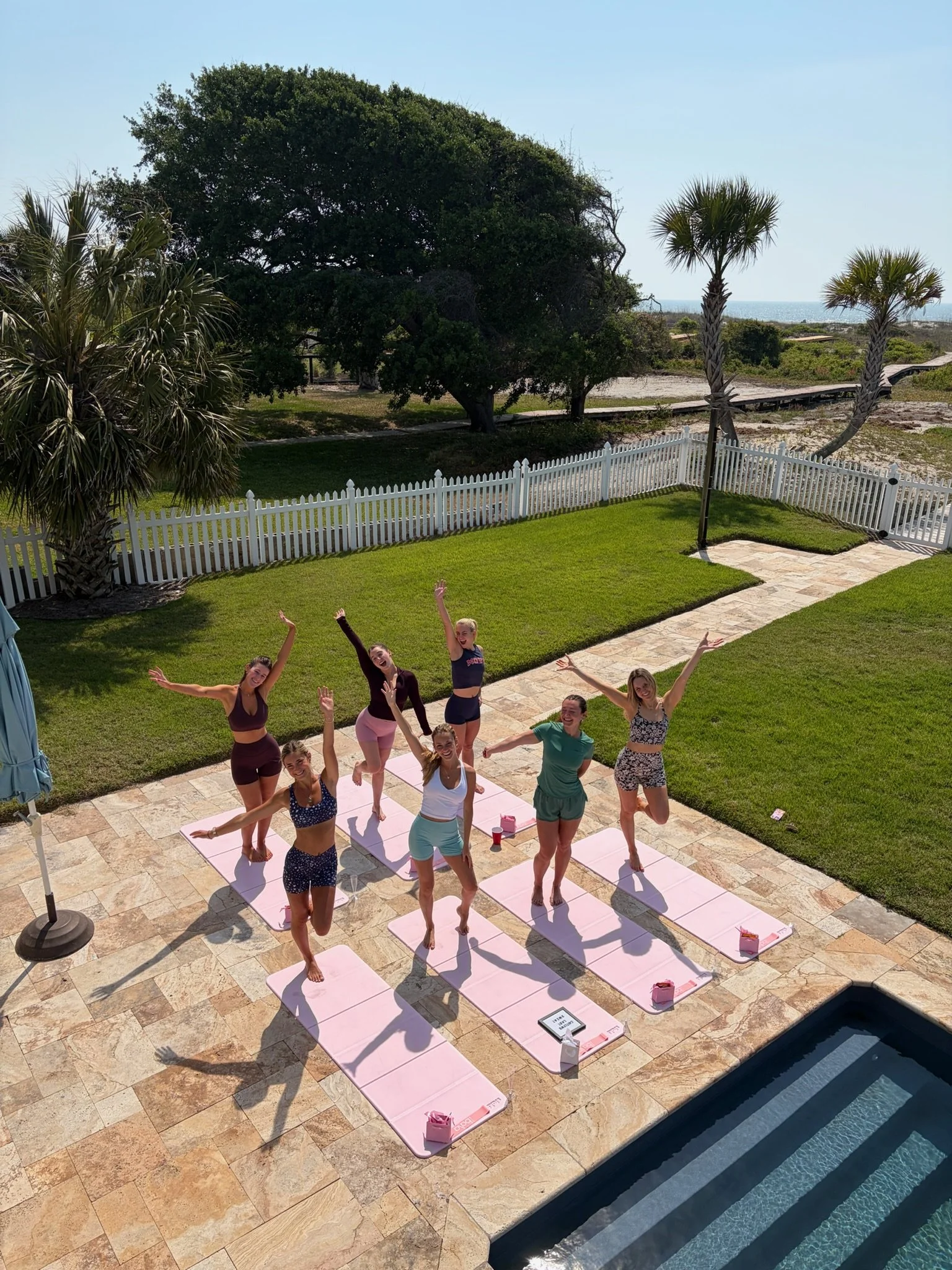 A group of women practicing yoga on pink mats outdoors near a swimming pool, with a white fencing, palm trees, large trees, and a view of the ocean in the background on a sunny day.