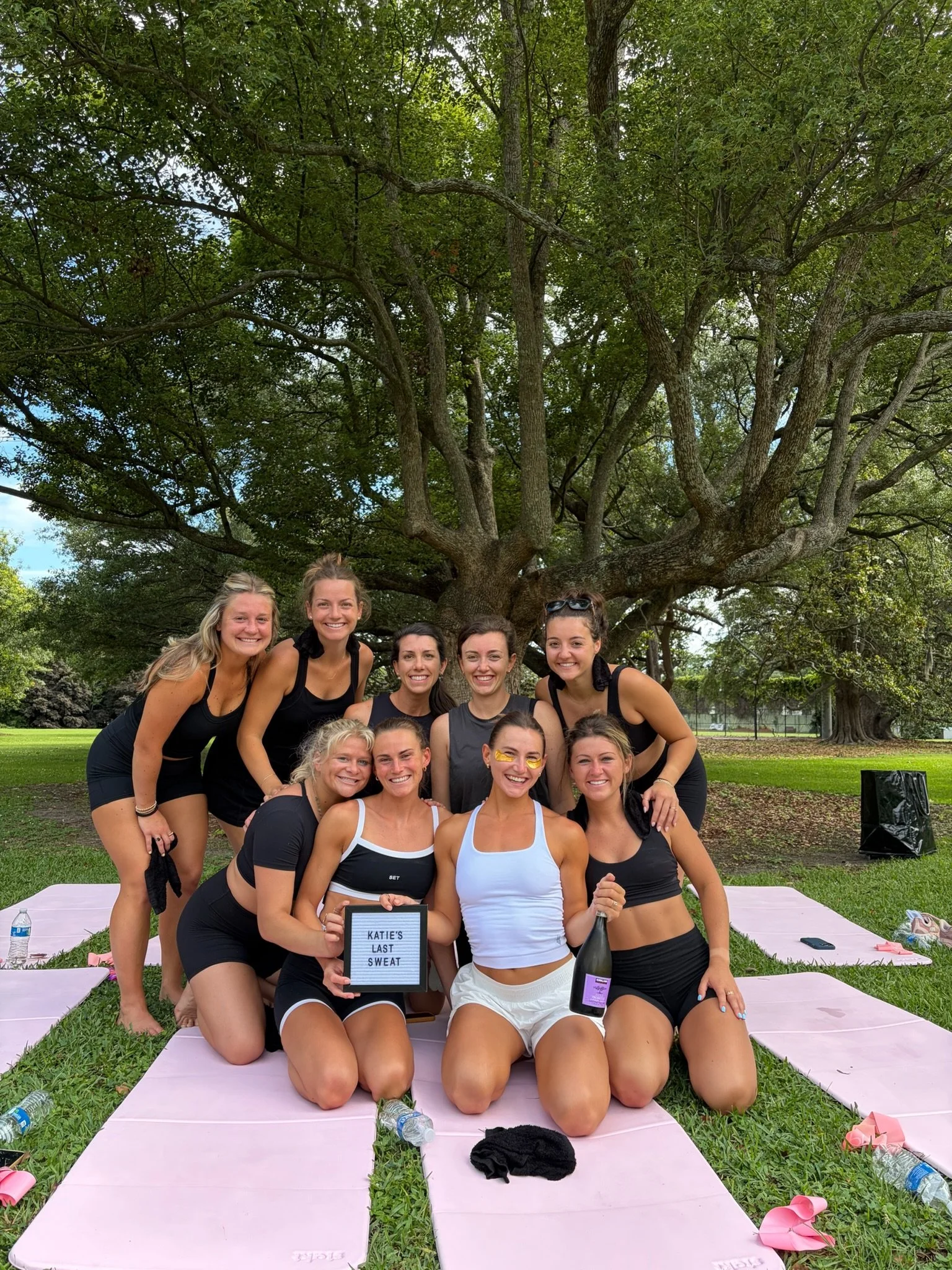 Group of women in athletic clothing posing outdoors under a large tree, some kneeling and some standing, with pink yoga mats on the grass, water bottles, and a sign that reads "KATIE'S LAST SWEAT" in front of them.