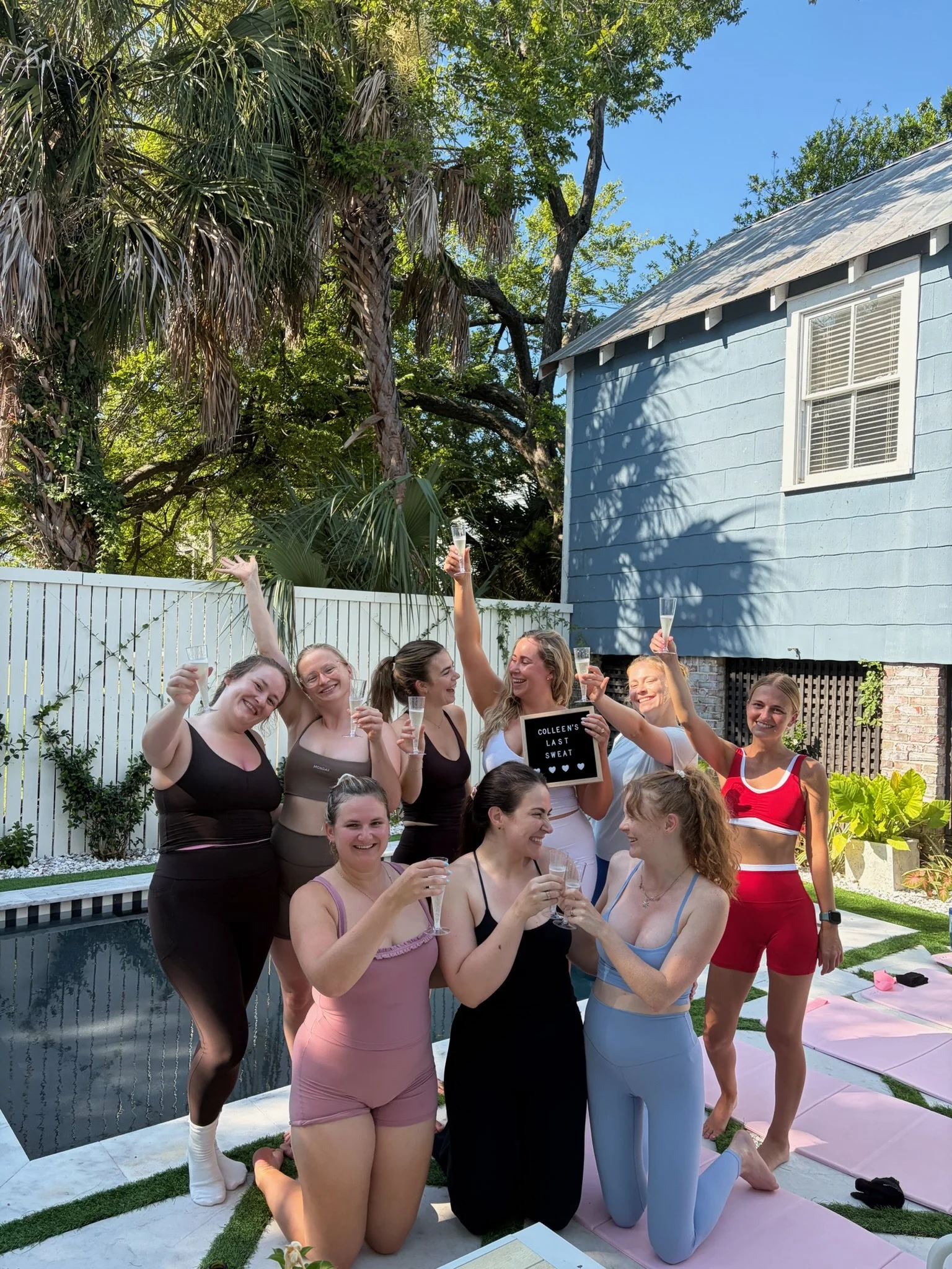 A group of women gather outdoors next to a pool, dressed in athletic or leisure clothing, some holding champagne flutes, celebrating together, with a house and lush green trees in the background on a sunny day.
