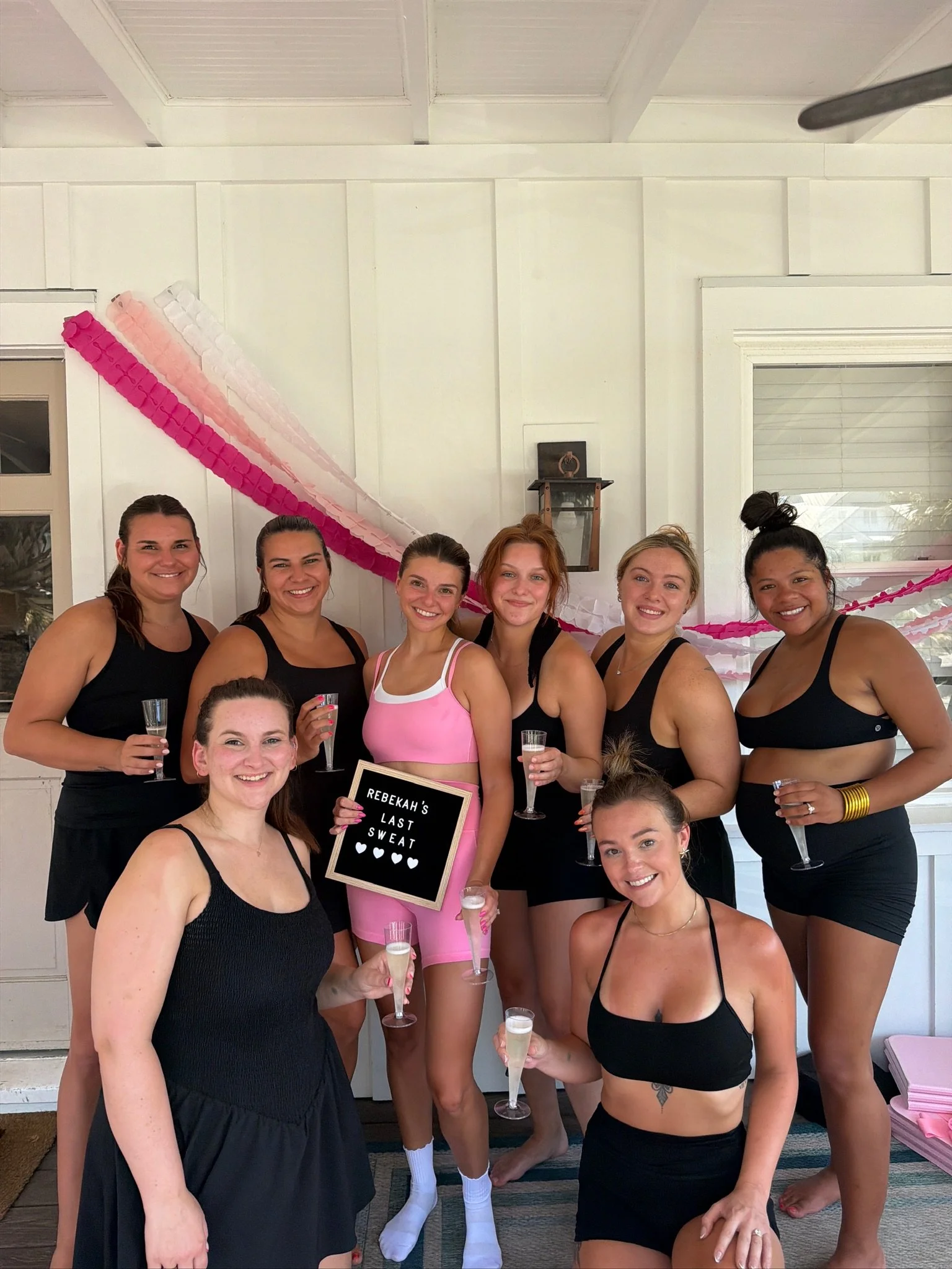 A group of nine women in athletic wear celebrating together at a party, holding champagne glasses, with pink and white decorations in the background.