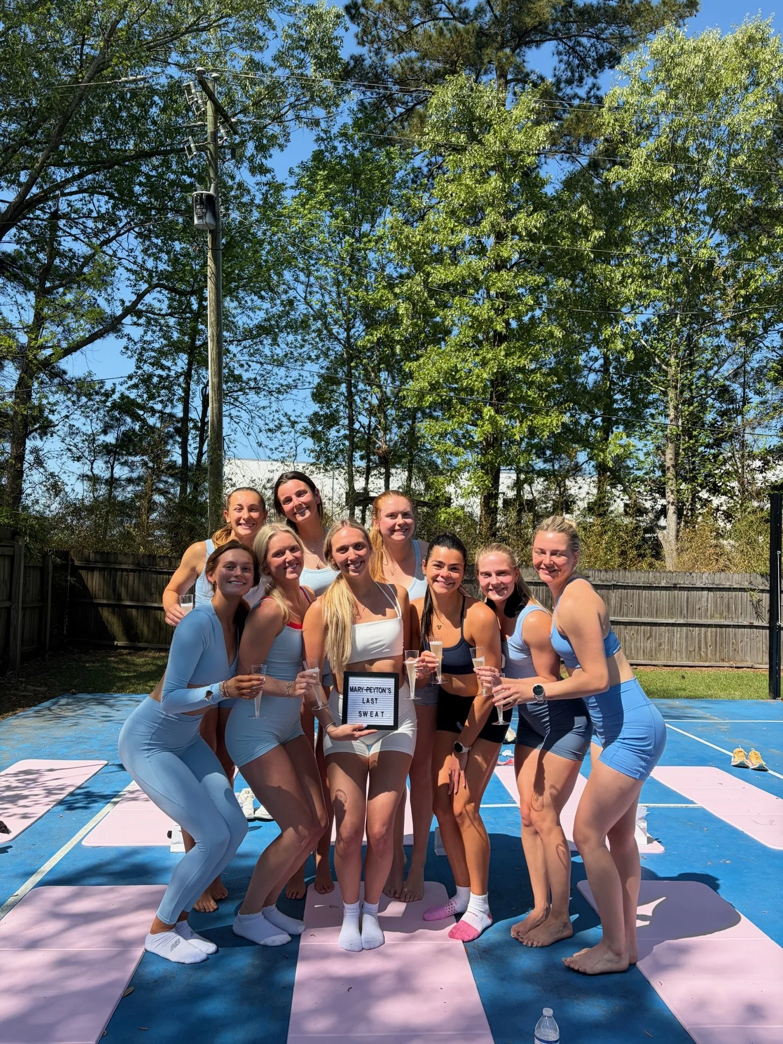 A group of women dressed in athletic wear holding glasses of champagne, standing together on a yoga mat outdoors, with trees and clear blue sky in the background. They are smiling and celebrating, with some holding a sign that reads "Mary-Peyton's Last Sweat."