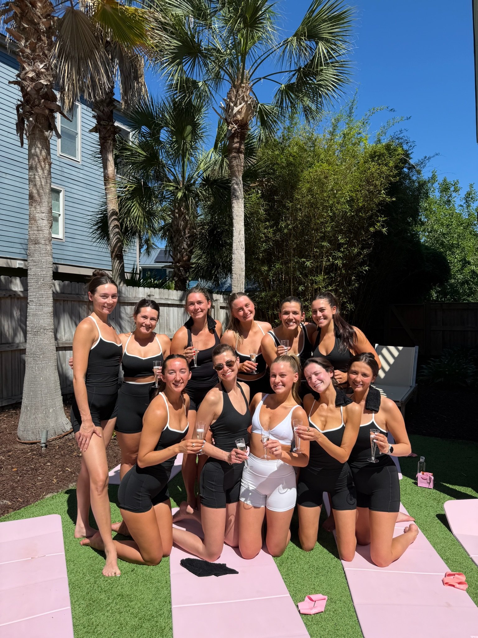 A group of ten women wearing black and white athletic outfits, holding glasses, and posing outdoors on pink yoga mats.