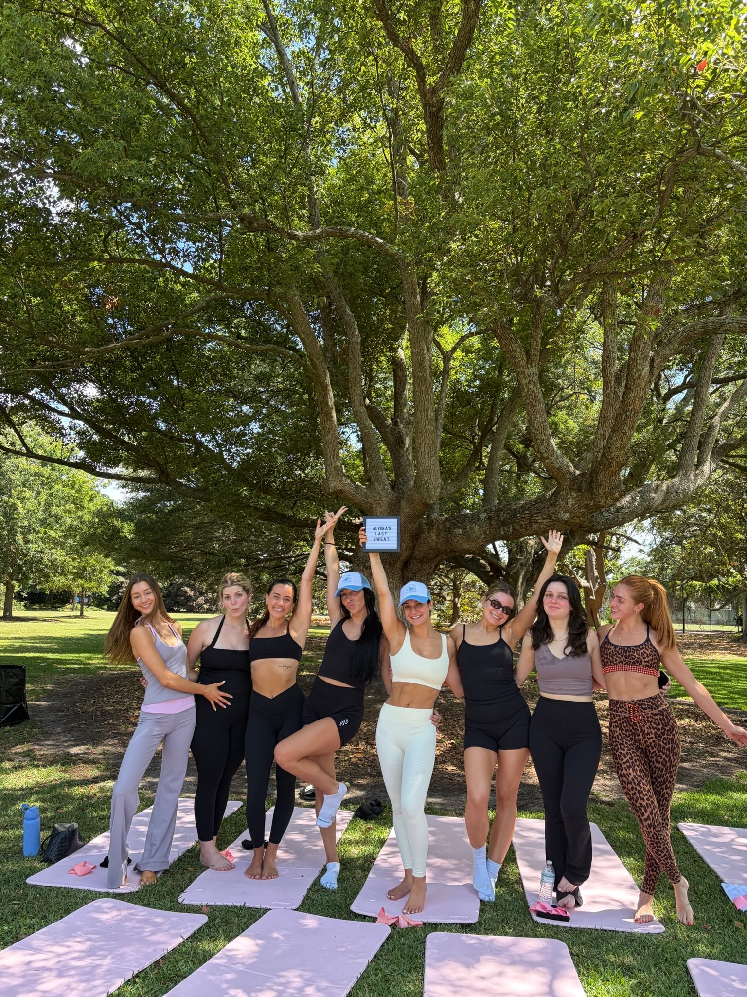 A group of women in workout clothes standing on yoga mats outdoors under a large tree, practicing yoga or stretching, with sunlight filtering through the leaves.