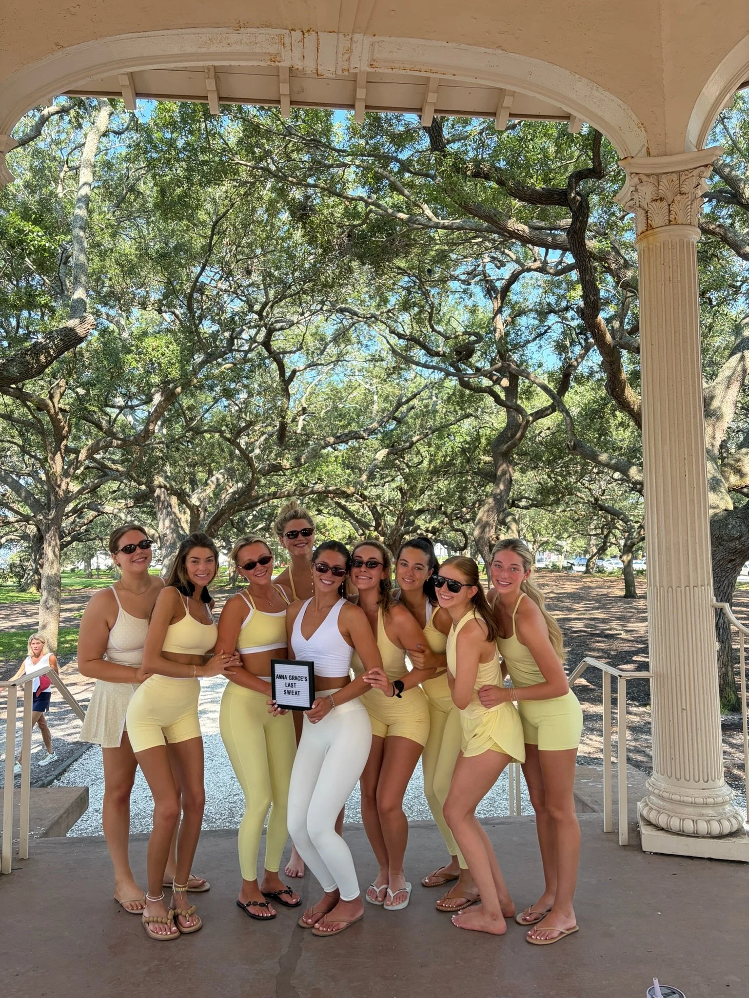 A group of ten women wearing yellow outfits, standing together under a large, arching porch with trees in the background, posing for a photo. One woman is holding a sign that reads 'Anna Grace's Last Sweat.'