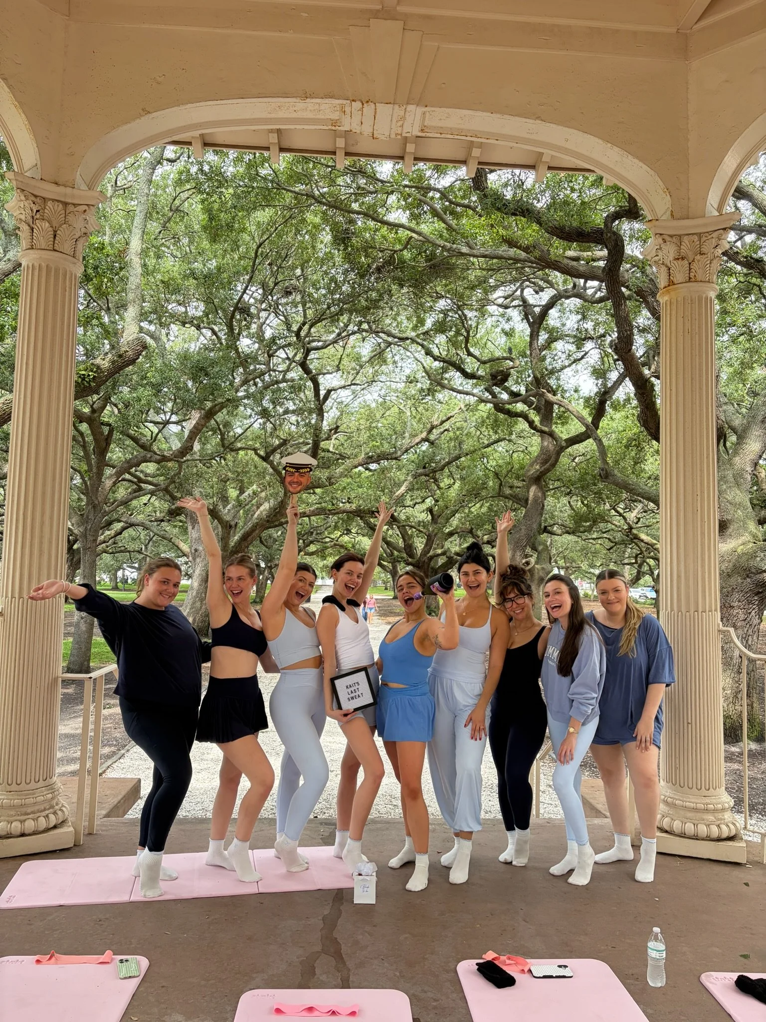 A group of women standing on a pink yoga mat inside a historic gazebo with columns, surrounded by large trees. They are dressed in workout clothes and smiling, celebrating a fitness achievement.
