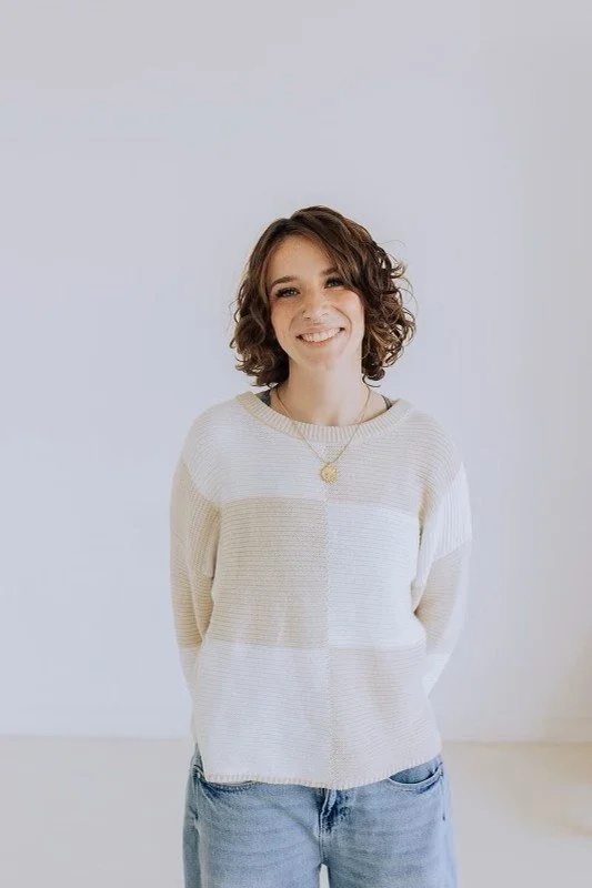 A smiling young woman with curly brown hair, wearing a white sweater and light blue jeans, standing in front of a plain light gray background.