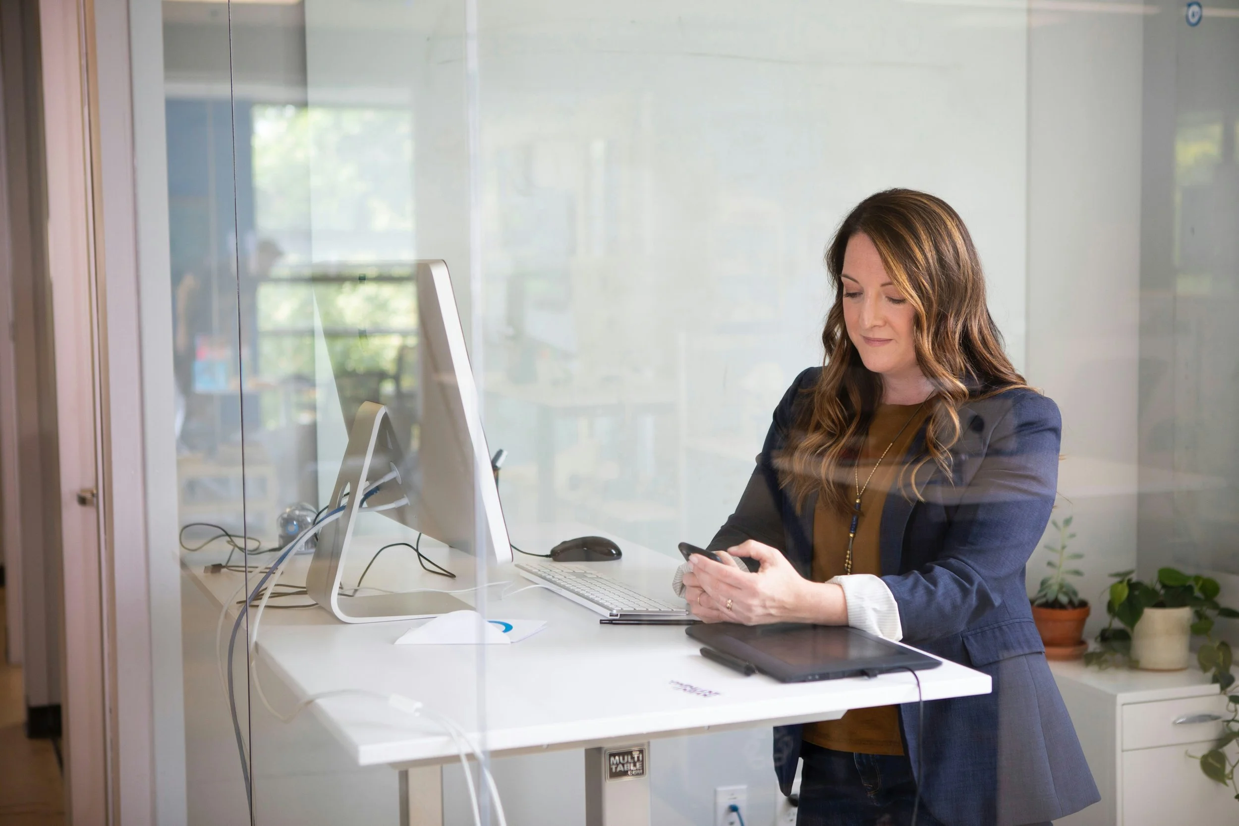 Woman in business attire standing in an office, looking at her phone, with a computer and plants in the background.