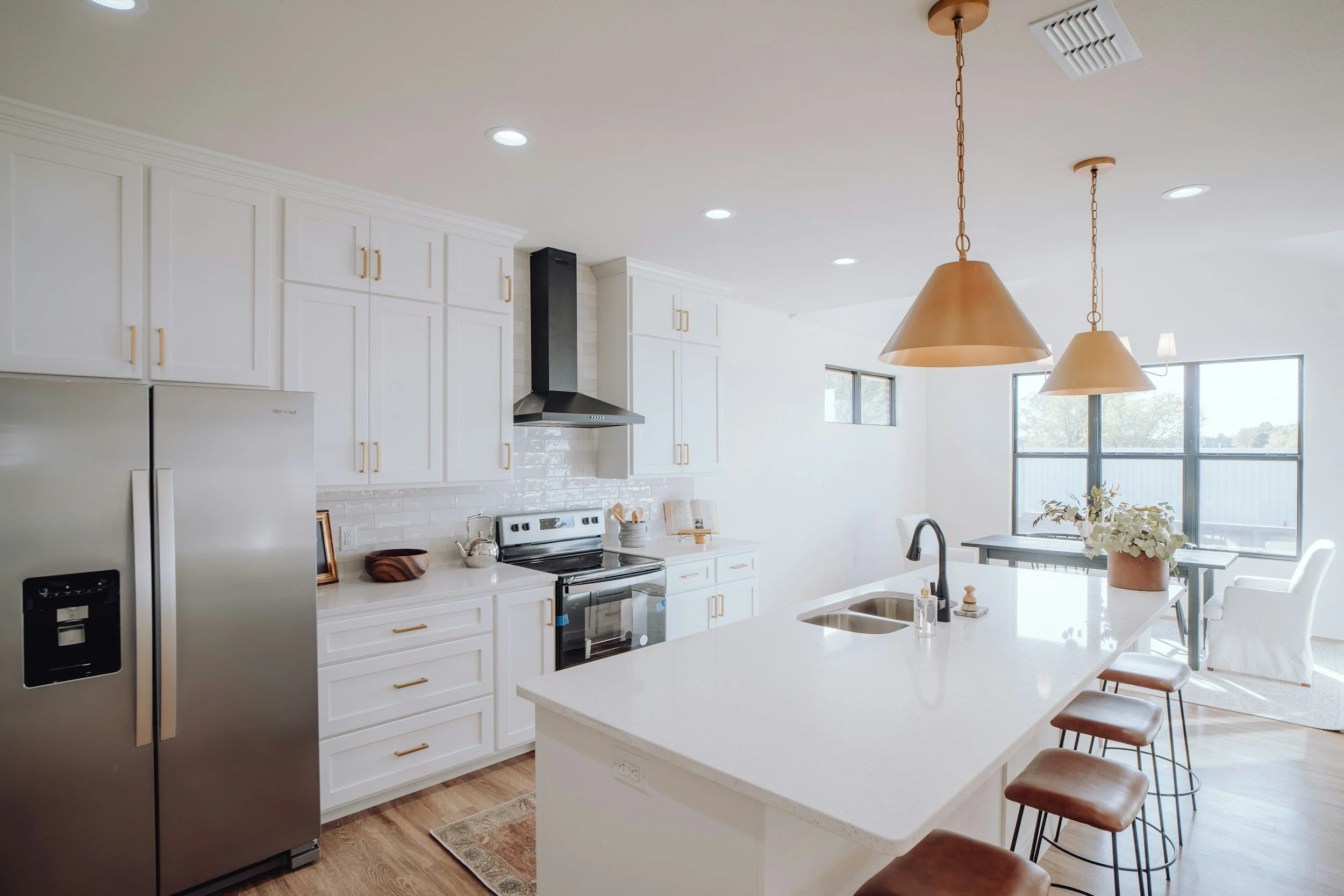 A modern kitchen with white cabinets, a stainless steel refrigerator, a black range hood, a white countertop island with a sink, and three hanging pendant lights. There are large windows in the background letting in natural light, a dining table with chairs, and decorative plants.