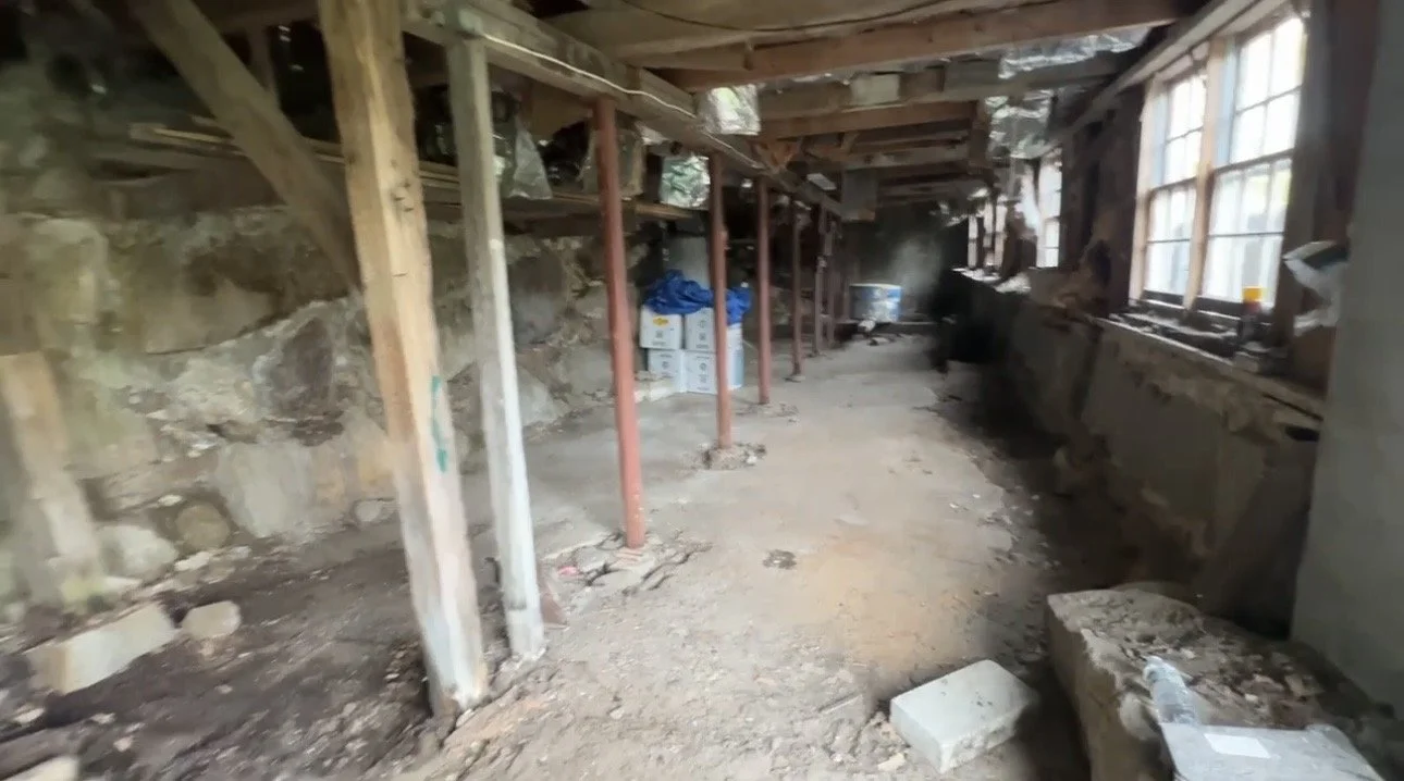 Construction site with exposed stone wall, wooden beams, scaffolding, and building supplies like boxes and tarps, near a window.
