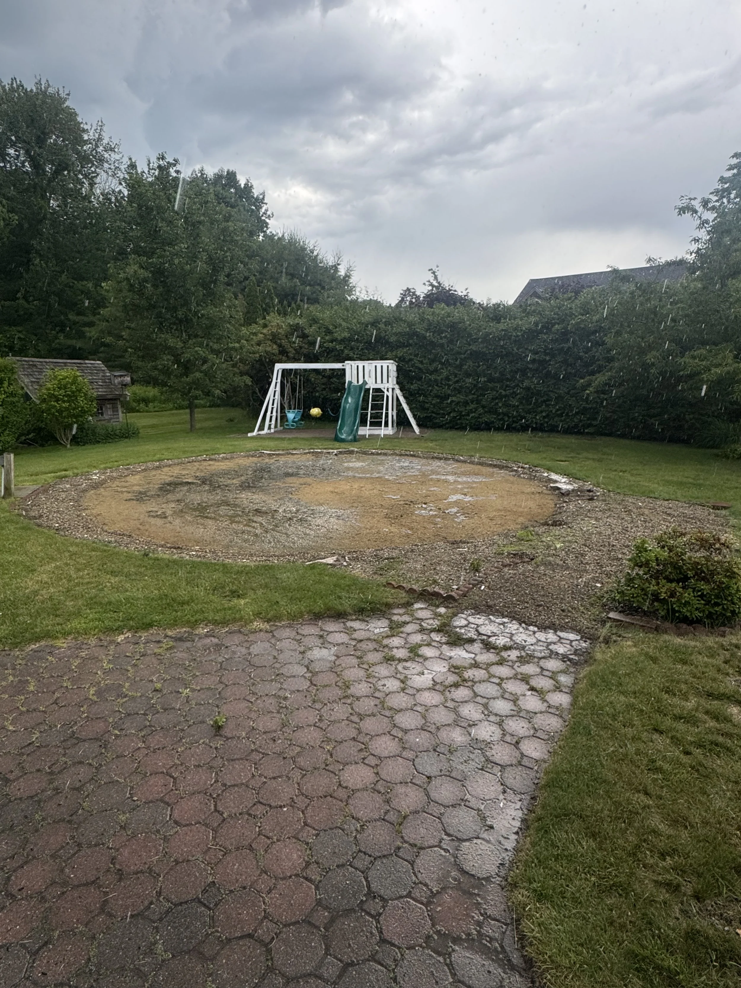 A backyard with a swing set including a slide and a small yellow object, surrounded by lush green trees, with dark storm clouds and rain overhead, and a partly cloudy sky.