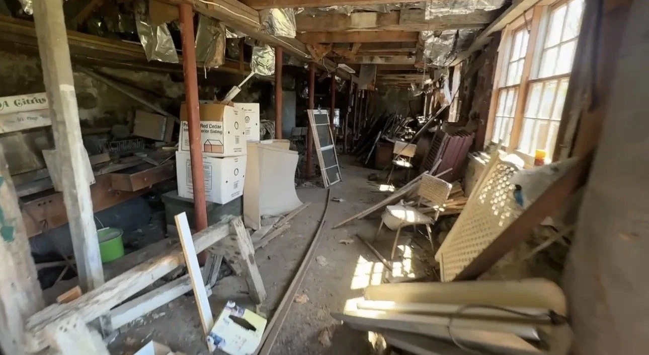 cluttered, messy attic filled with debris, old furniture, and cardboard boxes and other miscellaneous items piled up haphazardly, sunlight streaming through the windows.