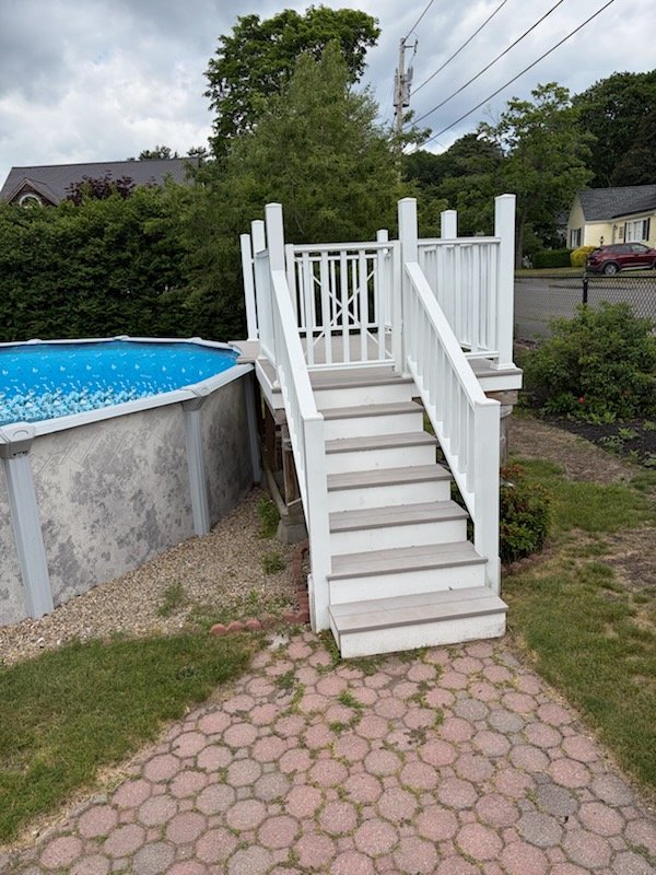 White wooden staircase leading up to a small deck with railing, next to an above-ground swimming pool, in a backyard with brick pathway, grass, bushes, and trees.