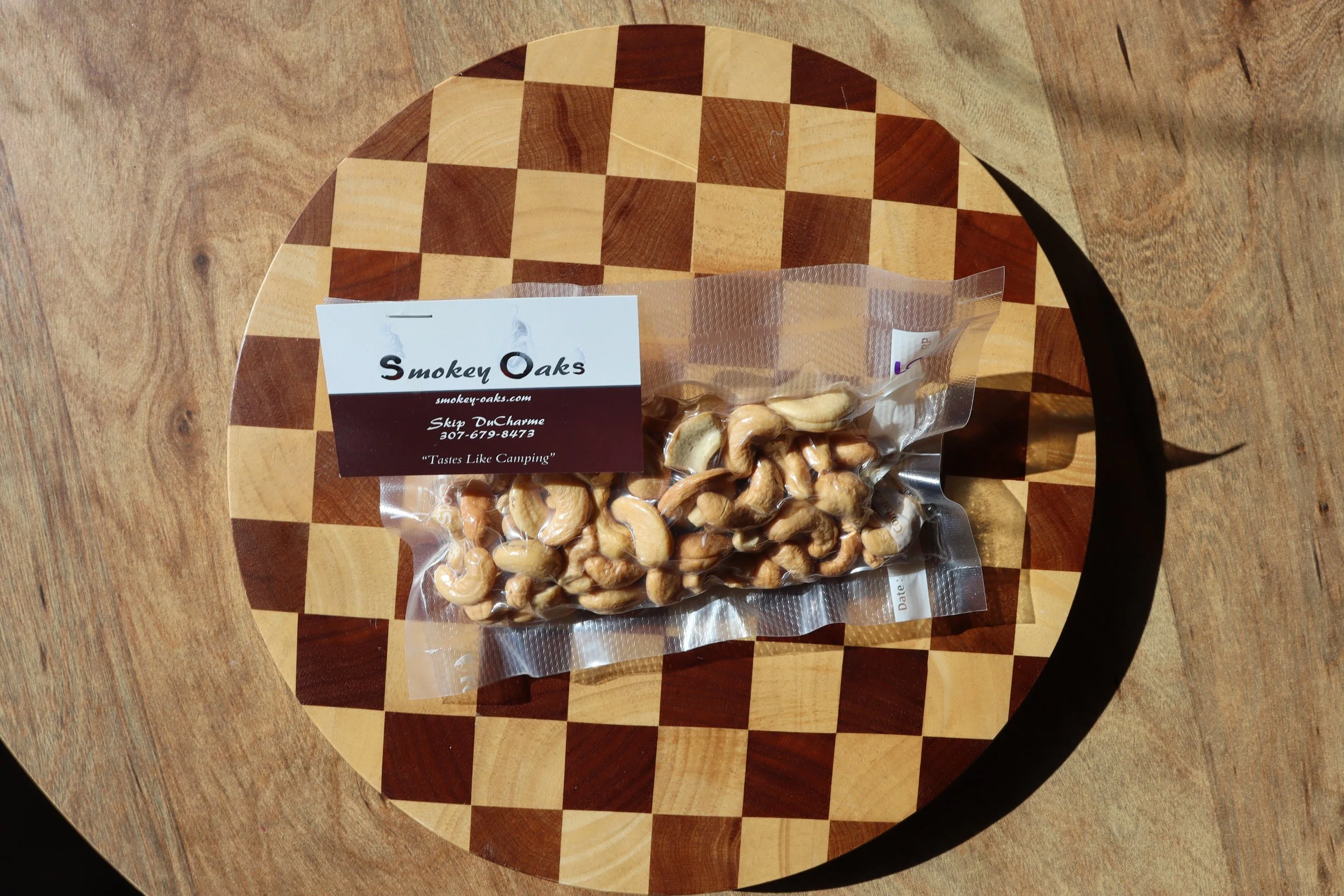 A plastic bag of shelled cashew nuts on a checkered wooden round cutting board placed on a wooden table.