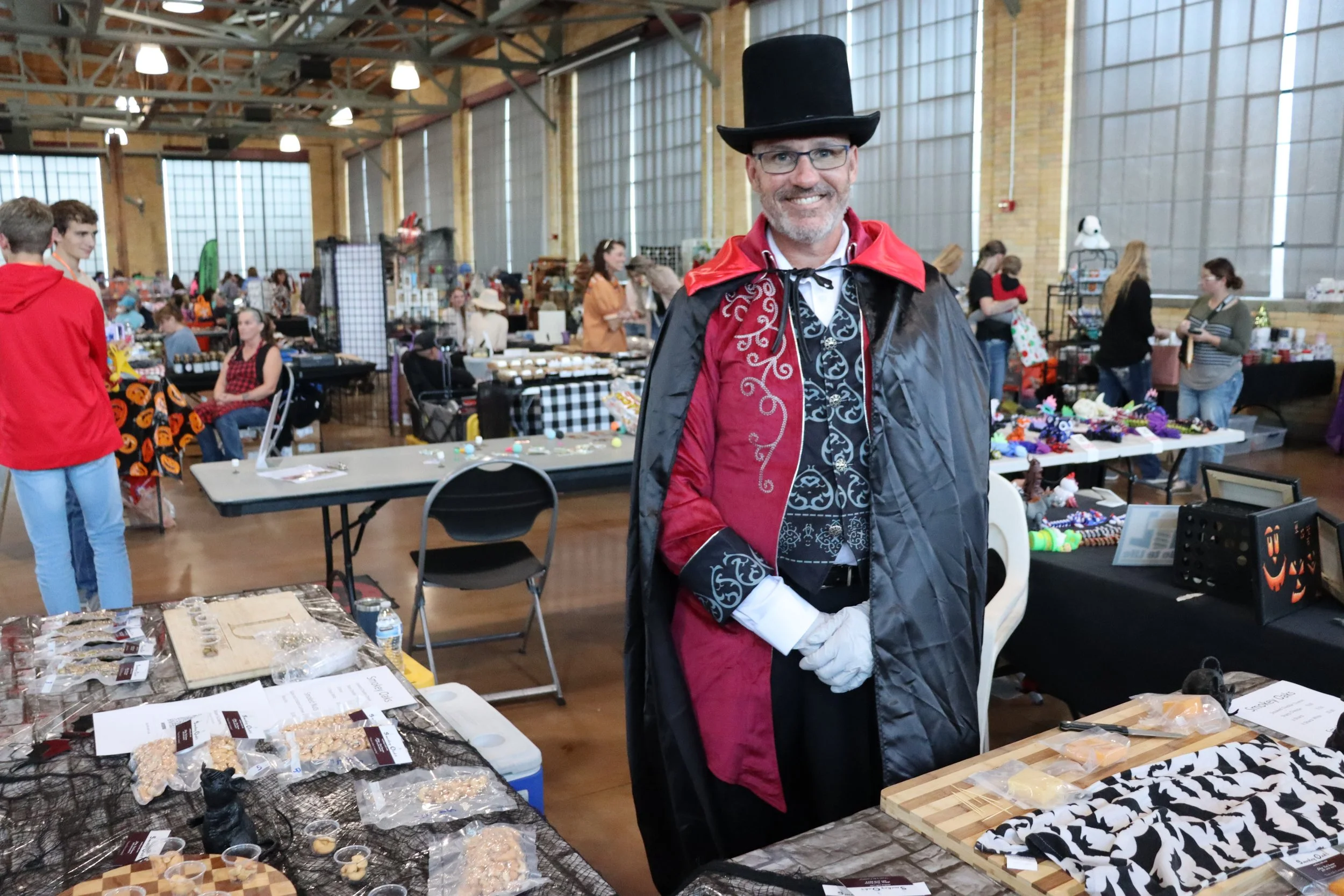 Man dressed as a Victorian vampire at a craft fair or market, surrounded by various handmade items on tables.