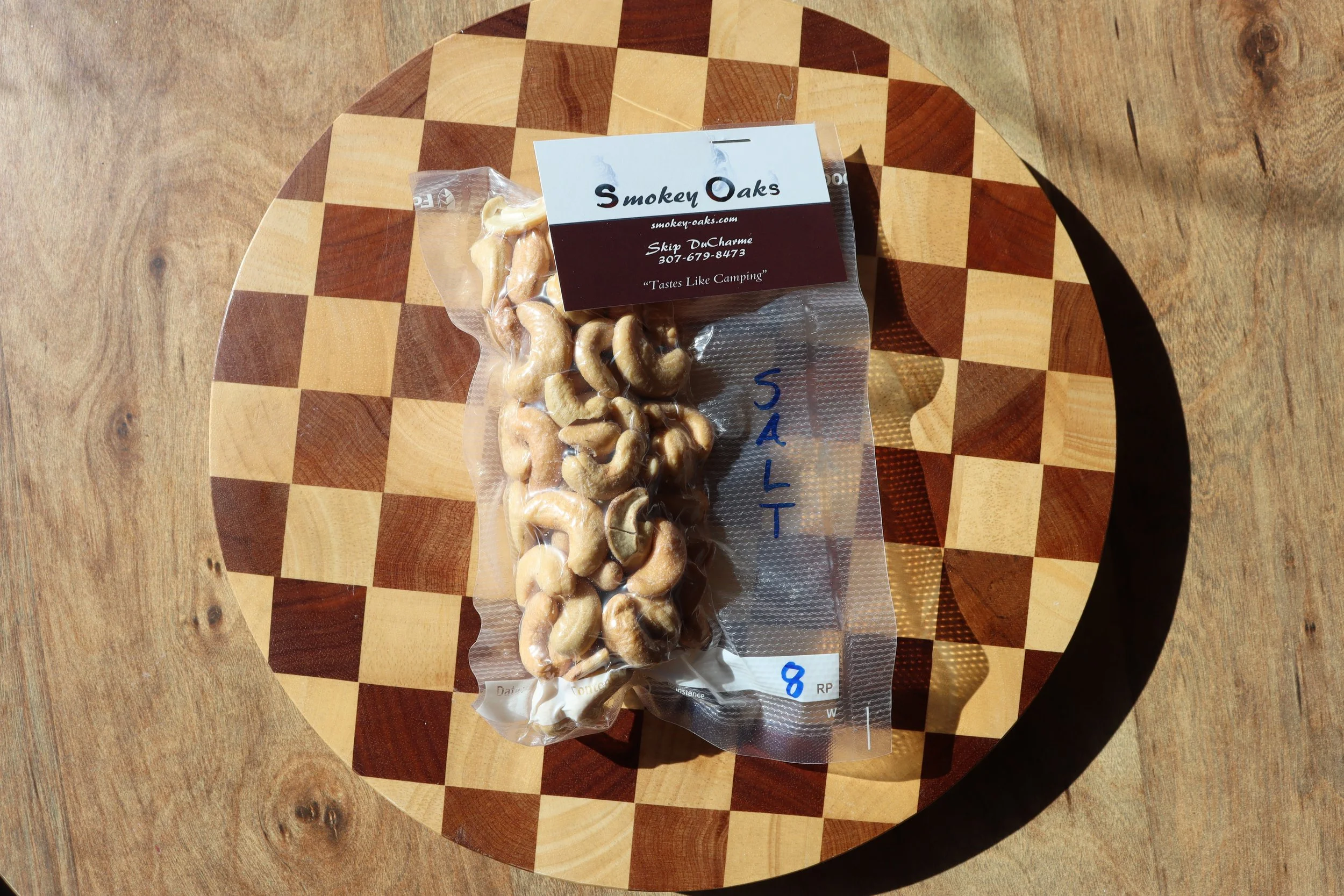 A plastic bag of salted cashews placed on a round wooden chessboard with alternating light and dark squares. The bag has a label from Smoky Oaks with contact information and the phrase 'Tastes Like Camping'.