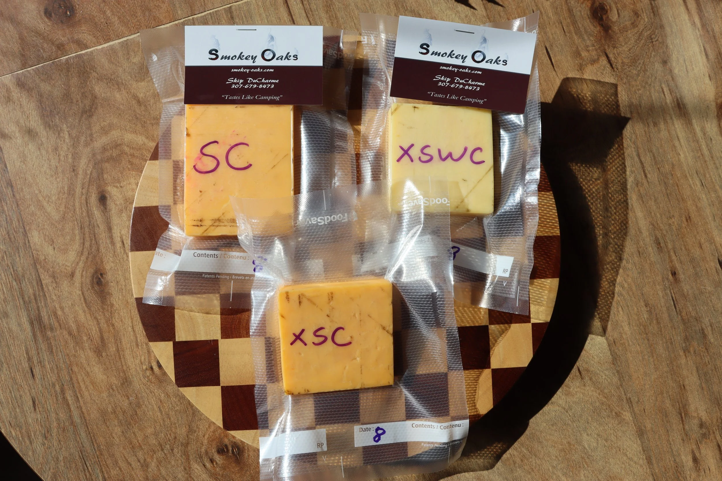 Three blocks of cheese in vacuum-sealed packaging on a wooden cutting board. The packaging has labels with handwritten markings: most say 'XSC' in purple ink, and two also have 'XSWC'. The cheese blocks are yellow with small brownish inclusions.