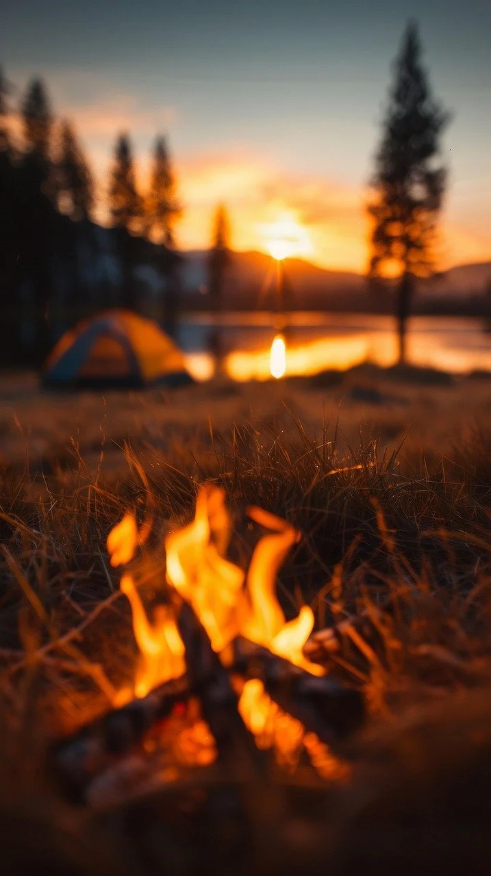 Campfire in the foreground with tents and a lake during sunset in the background, trees and mountains surrounding the scene.
