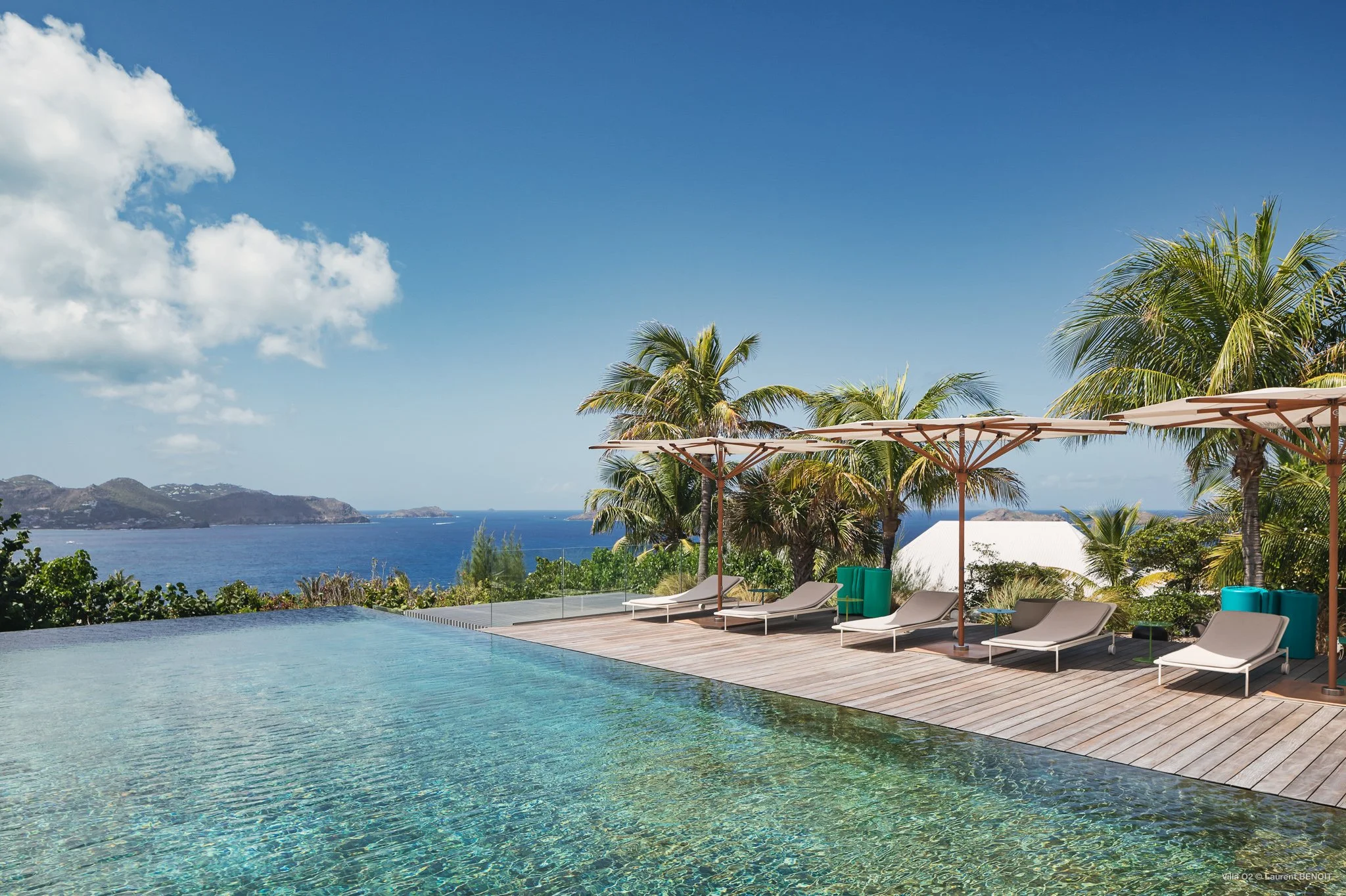 A woman relaxing on a lounge chair by an infinity pool with beach umbrellas, palm trees, and a view of the water and hills in the background